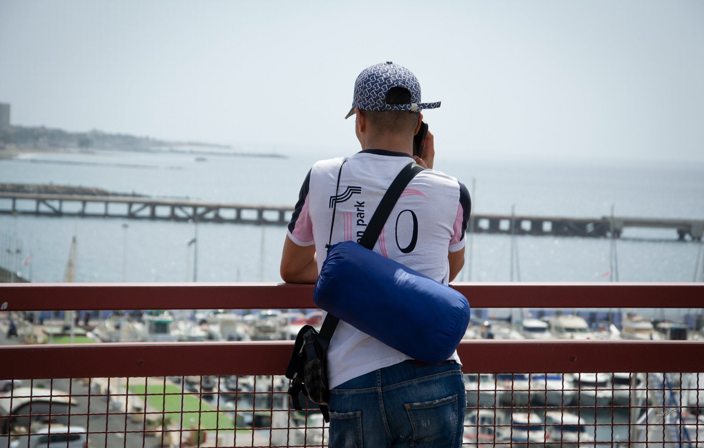 Dans cette image, un homme se tient de dos, regardant au loin vers la mer. Il est vêtu d'un t-shirt blanc avec des accents colorés et porte une casquette à motifs. Sur son épaule, il a un petit sac bleu. L'arrière-plan montre un port avec des bateaux amarrés et une jetée qui s'étend dans l'eau, créant une atmosphère tranquille. Le ciel est clair, suggérant une journée ensoleillée. L'homme semble concentré sur ce qu'il observe, profitant de la vue sur l'horizon maritime.