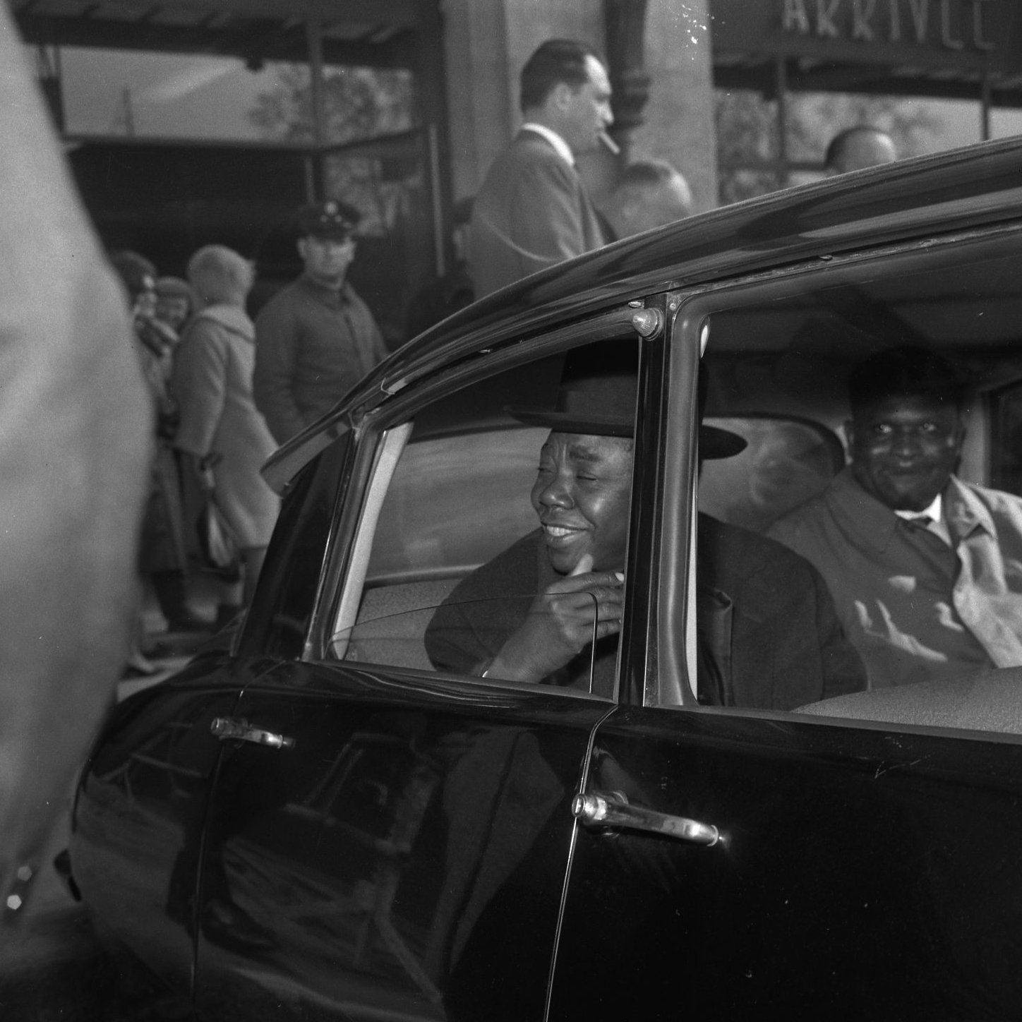 L'image montre l'intérieur d'une voiture noire, où deux hommes sont assis. L'un d'eux, qui se trouve à l'avant, semble souriant et engageant, habillé d'un costume élégant et portant un chapeau. Son expression indique qu'il est en train de parler ou de rire. À l'arrière de la voiture, l'autre homme a une expression plus neutre, vêtu également d'un manteau. À l'extérieur de la voiture, on peut apercevoir une foule de personnes en arrière-plan, certaines s'attardant dans un hall ou une gare d'arrivée. L'atmosphère semble animée, suggérant un moment de retrouvailles ou d'attente.
