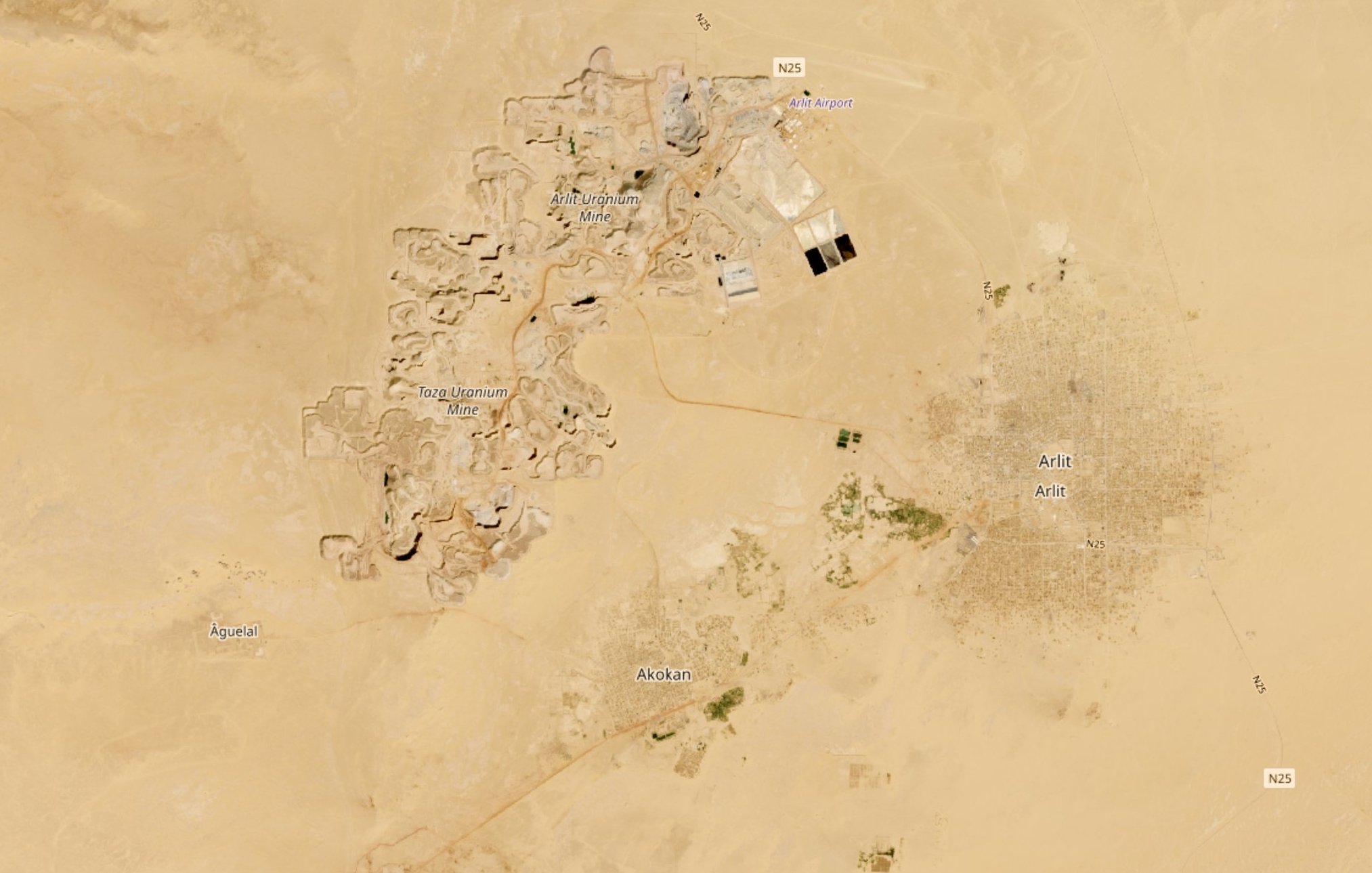 The image appears to be an aerial view of a desert landscape featuring two distinct areas. On the left side, there are ruins or archaeological sites, indicated by clusters of structures, possibly remnants of ancient buildings. Nearby, there is an airport and some roads connecting the areas. On the right side, there is a more densely populated area, showing what looks like a town or settlement named "Antic," characterized by a grid-like pattern, suggesting regular streets and organized layout. Surrounding the settlement, there are patches of greenery, likely indicating agricultural activity. The vast surrounding landscape is primarily sandy and barren, typical of desert regions.