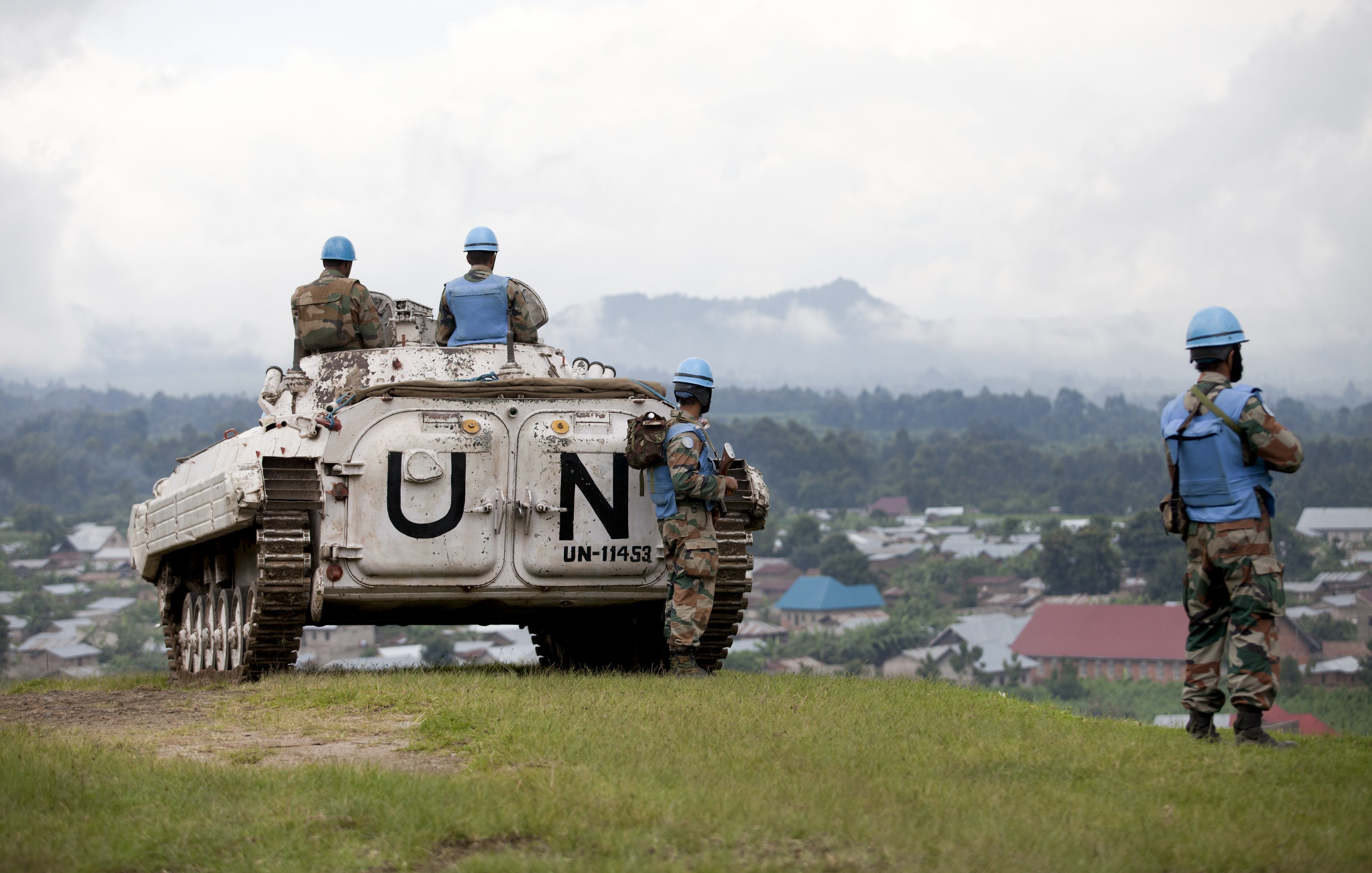 The image depicts a scene from a military operation involving United Nations peacekeepers. In the foreground, a military vehicle marked with "UN" is parked on a grassy area overlooking a landscape. Several soldiers in blue helmets, which signify their affiliation with the UN, are positioned around the vehicle. They appear to be surveying the area, which includes a view of a village with rooftops in the background. The atmosphere seems calm, with a cloudy sky adding to the setting. The soldiers' uniforms suggest they are part of a peacekeeping mission, emphasizing their role in maintaining stability in the region.