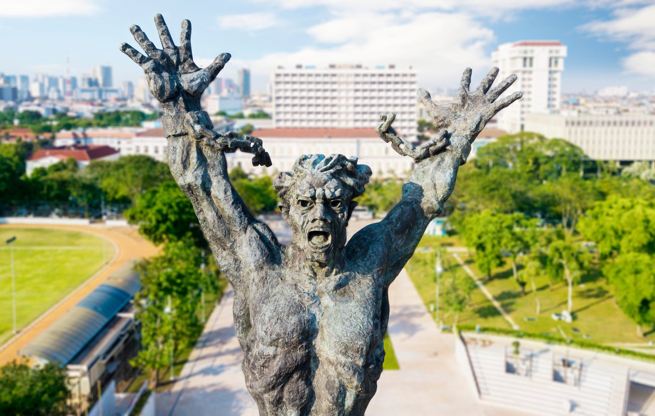 Cette image montre une statue impressionnante d'un homme, représenté dans une pose expressive. Il lève les bras vers le ciel, illustrant un sentiment intense, qui pourrait être celui de la lutte ou de l'angoisse. Son visage est déformé par une expression de détresse, accentuant une impression dramatique. La statue est en bronze, avec une texture riche et des détails finement sculptés qui soulignent les muscles et les traits faciaux. En arrière-plan, on aperçoit une ville moderne avec des bâtiments élevés et des espaces verts, créant un contraste entre la puissance de la sculpture et l'urbanisme environnant. Le ciel est dégagé, ajoutant une luminosité à la scène.