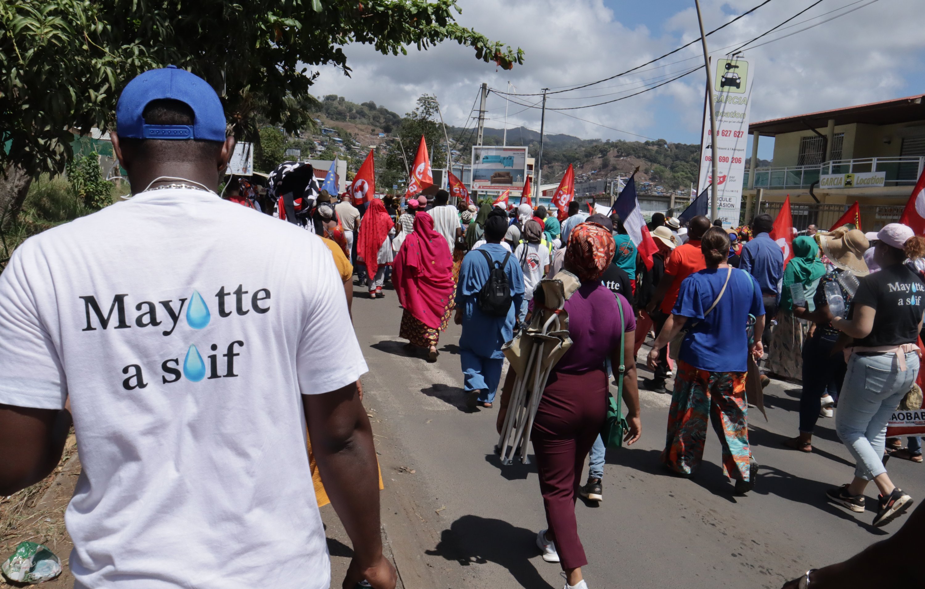 La scène dépeint un rassemblement animé dans une rue, où un groupe de manifestants avance. Au premier plan, on aperçoit une personne portant un t-shirt blanc avec l'inscription "Mayotte à souff" sur le dos. Les manifestants, vêtus de couleurs variées, portent des drapeaux rouges et d'autres pancartes, créant une atmosphère de solidarité et de revendication. À l'arrière-plan, on voit des bâtiments et des montagnes, sous un ciel partiellement nuageux. Les sons de la foule, les discussions et les chants de protestation résonnent, ajoutant à l'énergie de l'événement.