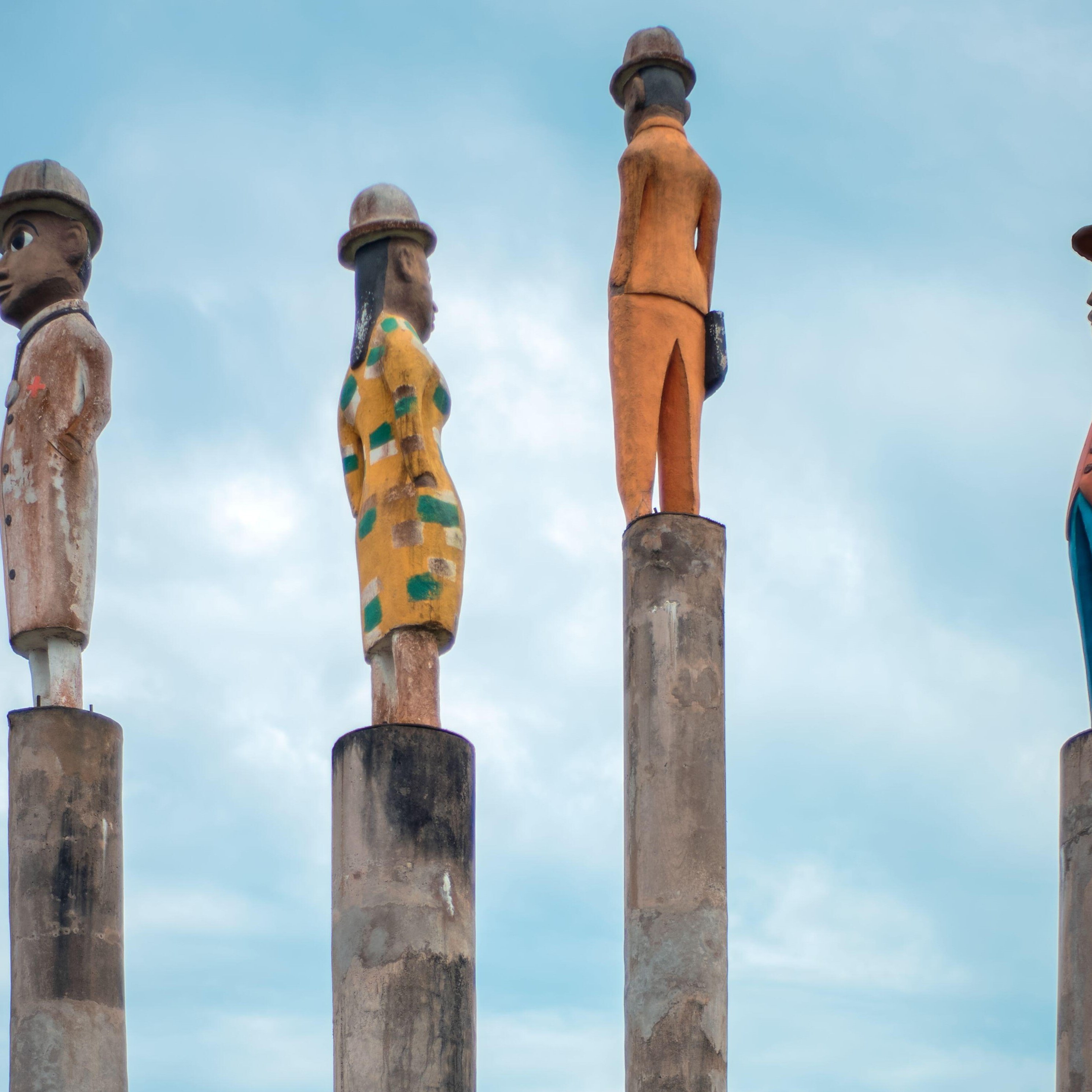 L'image présente quatre sculptures stylisées sur des colonnes. Chaque sculpture est peinte de manière différente, représentant des personnages humains : 1. La première sculpture à gauche est un homme avec un chapeau, vêtu d'un costume. Son expression est sérieuse. 2. La deuxième sculpture est une femme qui porte une robe avec un motif à rayures. Elle se tient droite, les bras le long du corps. 3. La troisième sculpture est un homme en orange, qui se tient également droit mais légèrement tourné sur le côté. 4. Enfin, la dernière sculpture est un homme en costume bleu, avec un chapeau, regardant vers l'horizon. L'arrière-plan est dominé par un ciel bleu clair, créant une ambiance paisible. Les sculptures semblent faire partie d'une installation artistique qui évoque la culture et l'identité. Les colonnes sur lesquelles elles reposent ajoutent une touche de hauteur et de grandeur à la scène.