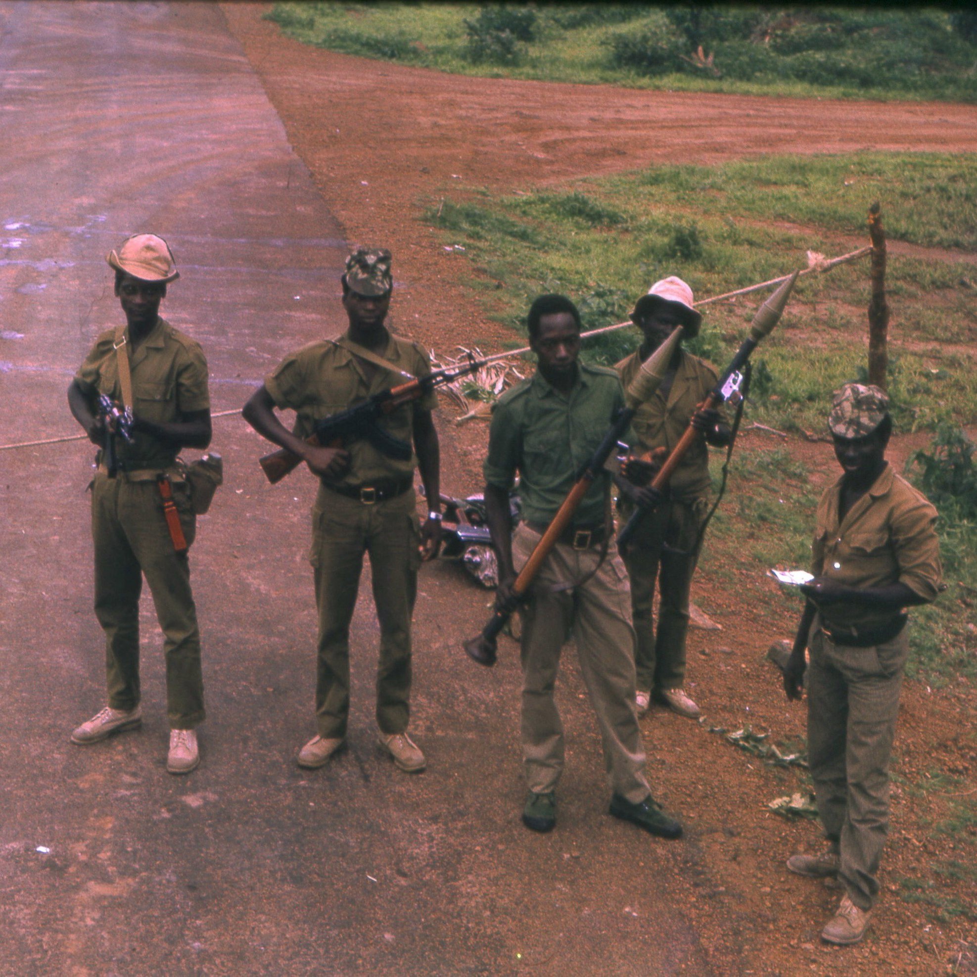L'image montre un groupe de cinq soldats en uniforme, debout sur une route non pavée. Ils portent des chapeaux, et chacun d'eux tient une arme à feu. Le sol est poussiéreux, avec quelques zones humides visibles. En arrière-plan, on voit une végétation luxuriante, typique d'un environnement tropical. Les soldats affichent des postures sérieuses, et certains d'entre eux paraissent concentrés sur leur environnement. Cette scène suggère un contexte de sécurité ou de patrouille.