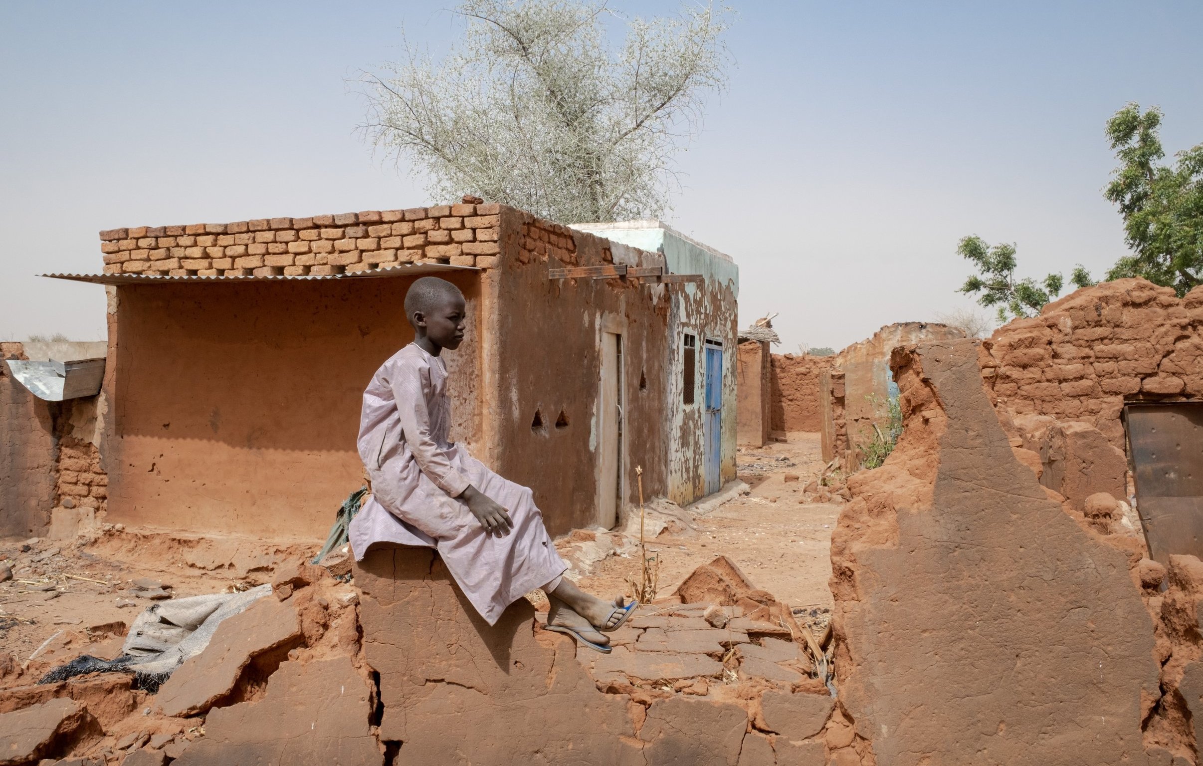 L'image montre un jeune garçon assis sur un mur de terre, au milieu d'un paysage rural aride. Il porte une longue tunique claire et semble pensif. En arrière-plan, on voit des maisons en terre battue, avec des murs qui sont fissurés ou partiellement détruits. L'environnement est sec, avec quelques arbres isolés. Le ciel est dégagé, d'un bleu clair. L'atmosphère générale transmet une sensation de solitude et de résilience dans un milieu rural difficile.
