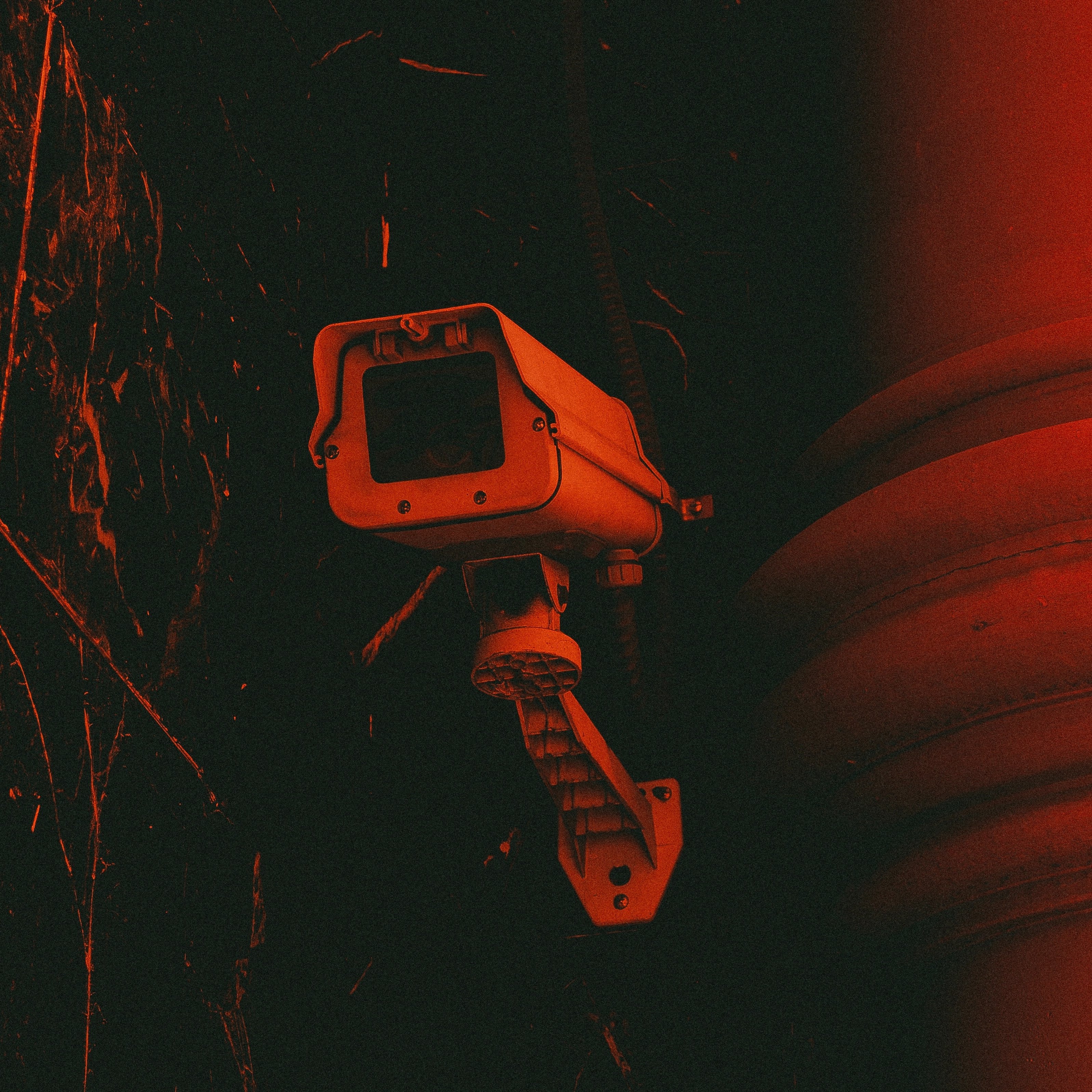 L'image représente une caméra de surveillance fixée à un mur. La caméra, avec son boîtier rectangulaire et son objectif, est peinte en gris clair. Elle est montée sur un support qui lui permet de pivoter, capturant ainsi une vaste zone de surveillance. L'arrière-plan est d'un rouge intense, créant une atmosphère sombre et légèrement inquiétante. Des textures sombres et des motifs abstraits se dessinent autour, ajoutant à l'effet dramatique de l'image. Cette combinaison de couleurs et de formes suggère une ambiance de vigilance et de sécurité.