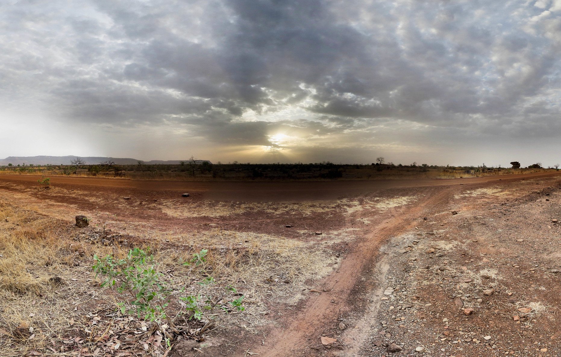 Imaginez un vaste paysage ouvert, dominé par des tons chauds de terre et des nuances de beige. Le sol est sec et parsemé de petites pierres, avec des herbes jaunies qui émergent de la terre aride. À l'horizon, un ciel nuageux s'étend, teinté de gris avec des rayons de lumière qui percent, annonçant un coucher de soleil. La lumière crépusculaire crée une ambiance paisible, tandis que la nature environnante semble calme et tranquille. Les silhouettes lointaines de collines ajoutent une dimension au paysage, renforçant le sentiment d'immensité et d'isolation.
