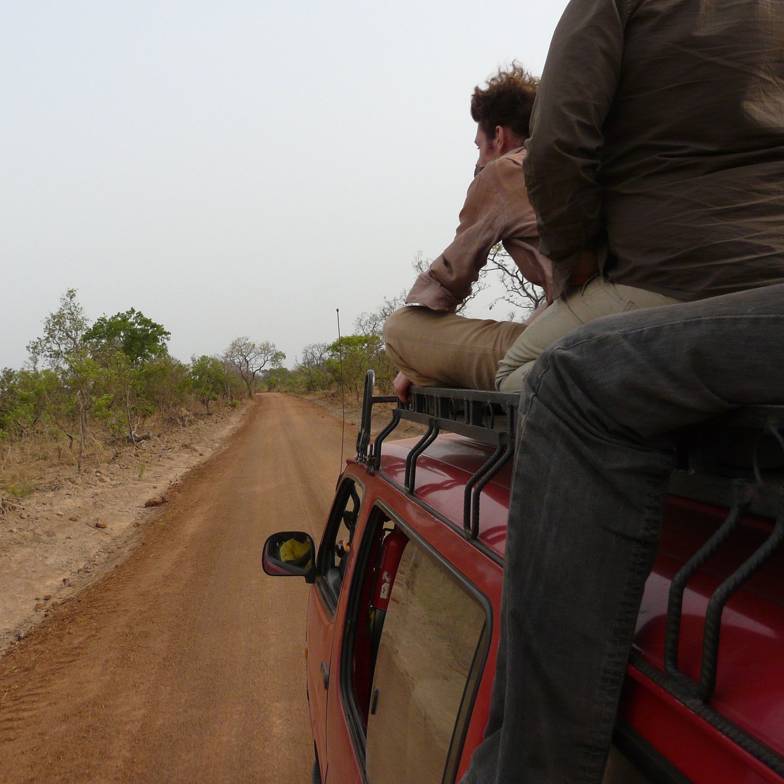 Imaginez un paysage d'arrière-pays, où un chemin de terre étroit s'étend devant vous. Sur les côtés, on peut voir de petites plantes et des buissons épars, témoignant d'une nature sauvagement préservée. La route est poussiéreuse et légèrement sinueuse, suggestive d'une aventure en milieu rural. Deux personnes sont assises sur le toit d'un véhicule, l'une d'elles se penche en arrière, le regard perdu au loin. La scène évoque un sentiment de liberté et d'exploration. L'air est probablement chargé de chaleur et du bruit léger de la nature environnante. Les couleurs sont chaudes, avec des teintes de rouge et de brun du sol, et un ciel lumineux qui crée une ambiance de voyage au cœur de la nature.