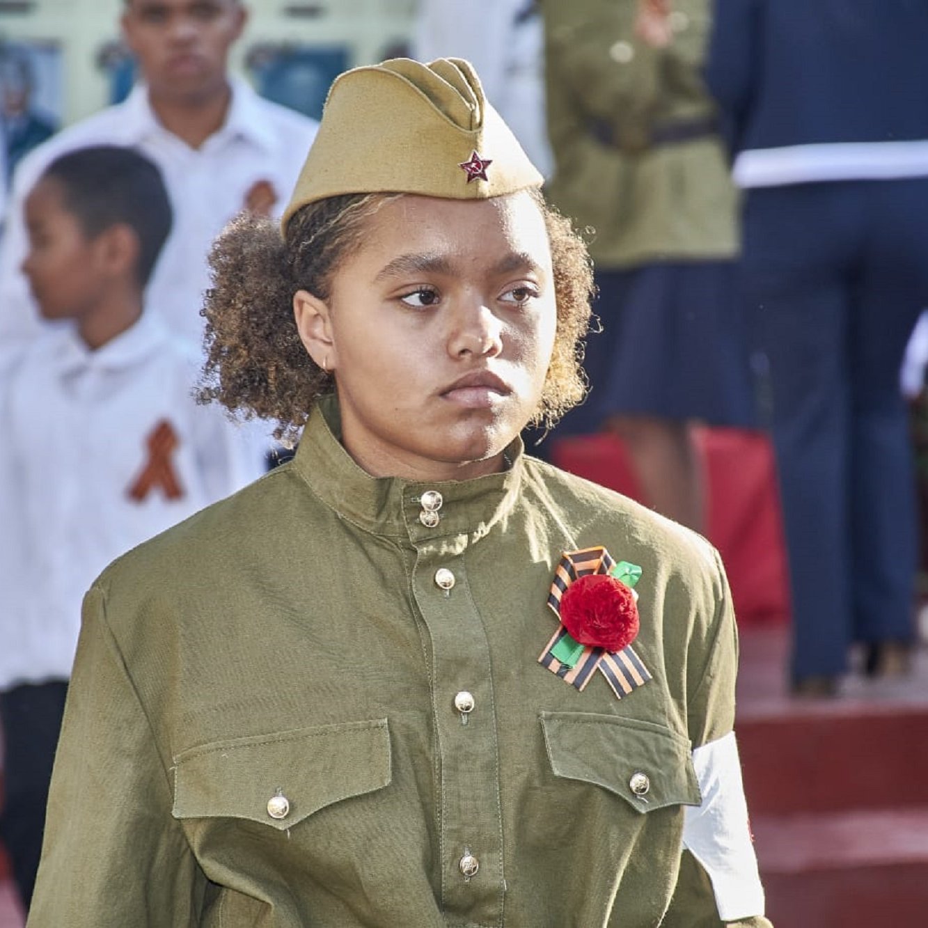 The image features a young girl wearing a military-style uniform with a khaki color. She has a military hat adorned with a star. The uniform includes decorative items like a red flower and a ribbon, possibly indicating a celebration or commemorative event. In the background, there are other children and adults dressed in formal attire, suggesting a public gathering or ceremony, likely related to a historical event. The setting appears to be outdoors, with a backdrop of steps and other participants.