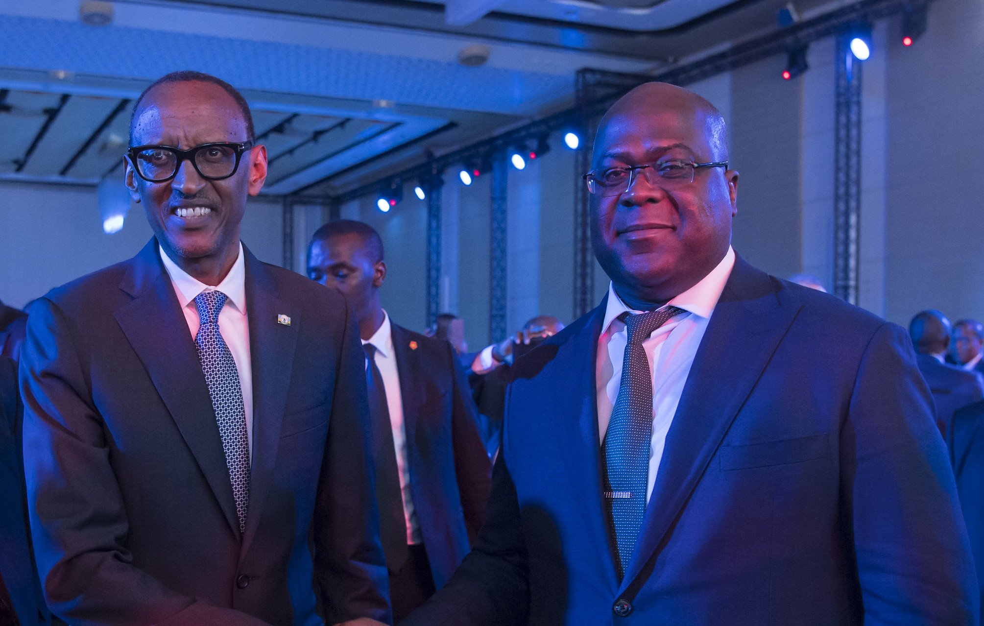 L'image montre deux hommes en costume qui se tiennent debout, se serrant la main dans un contexte de réunion ou d'événement officiel. L'un porte des lunettes et semble d'origine rwandaise, tandis que l'autre est un homme robuste, légèrement plus âgé, probablement d'origine congolaise. Ils affichent des expressions souriantes et amicales, dans une ambiance éclairée par des lumières bleues créant une atmosphère conviviale. En arrière-plan, on peut entrevoir d'autres personnes présentes à l'événement.