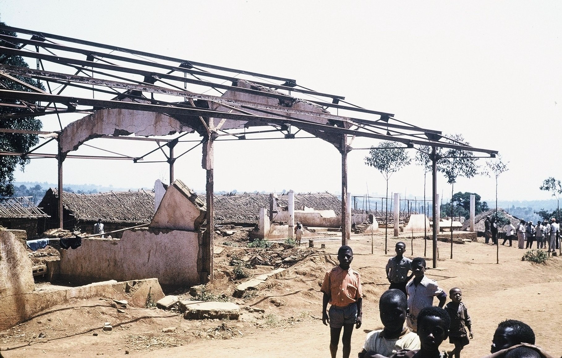 L'image montre un paysage de dévastation avec des structures en ruine. À l'avant-plan, plusieurs personnes, dont des enfants et des adultes, se tiennent debout sur un sol poussiéreux. En arrière-plan, on voit des bâtiments partiellement effondrés, avec des toits manquants et des murs endommagés. Des arbres isolés se dressent ici et là, ajoutant une touche de verdure à ce cadre désolé. Le ciel est dégagé et ensoleillé, contrastant avec l'état des lieux. L'atmosphère suggère une scène de désolation, peut-être après un événement tragique.