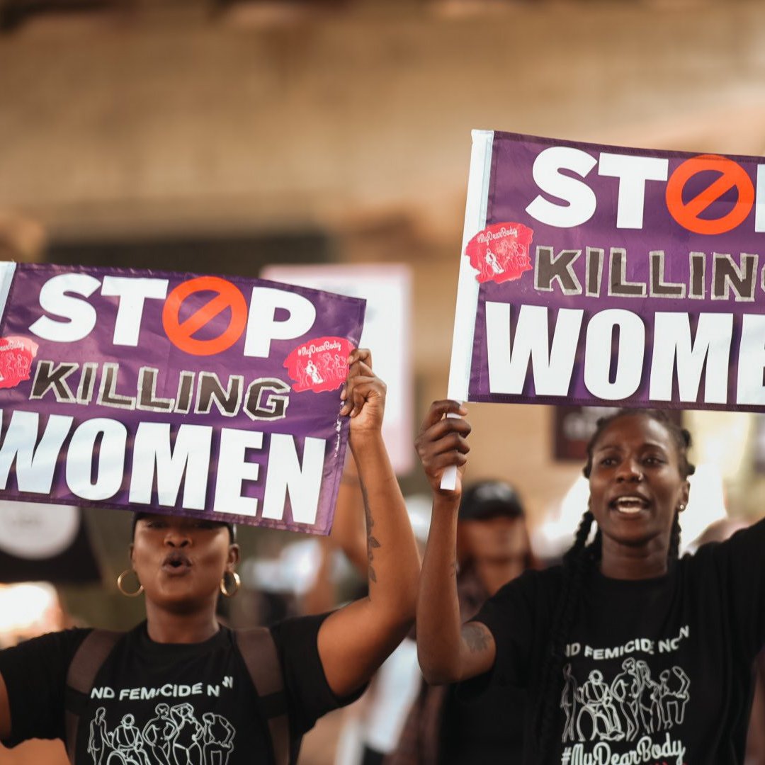 L'image montre deux femmes participante à une manifestation. Elles tiennent chacune une grande pancarte. Les pancartes portent le message "STOP KILLING WOMEN" en lettres capitales de couleur blanche, avec un symbole barré en rouge. Les femmes ont des expressions déterminées et sont vêtues de t-shirts noirs. L'arrière-plan suggère une ambiance de mobilisation avec d'autres personnes qui marchent et participent à l'événement. L'atmosphère est chargée d'émotion et de solidarité.