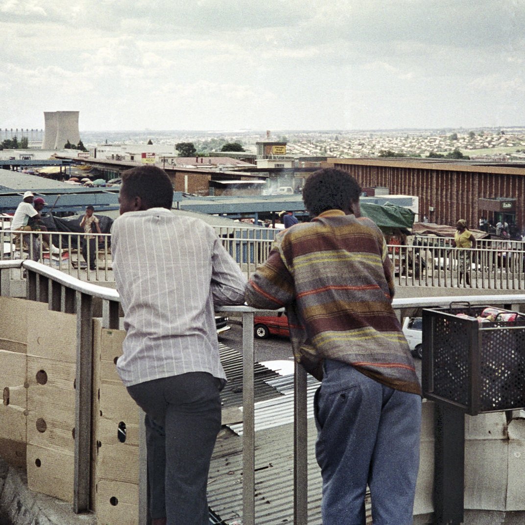 Dans cette image, on voit deux hommes debout sur un balcon surélevé, se penchant contre la balustrade. Ils regardent en bas un marché animé. Leurs silhouettes sont tournées vers l'extérieur, révélant un paysage urbain qui s'étend derrière eux, avec des bâtiments et des petites boutiques. L'environnement est vivant, rempli de personnes et d'activités. Le ciel est nuageux, ajoutant une ambiance mélancolique à la scène. Les vêtements des hommes suggèrent une certaine simplicité, avec des motifs discrets. L'ensemble dégage une atmosphère de contemplation et d'observation de la vie quotidienne en ville.