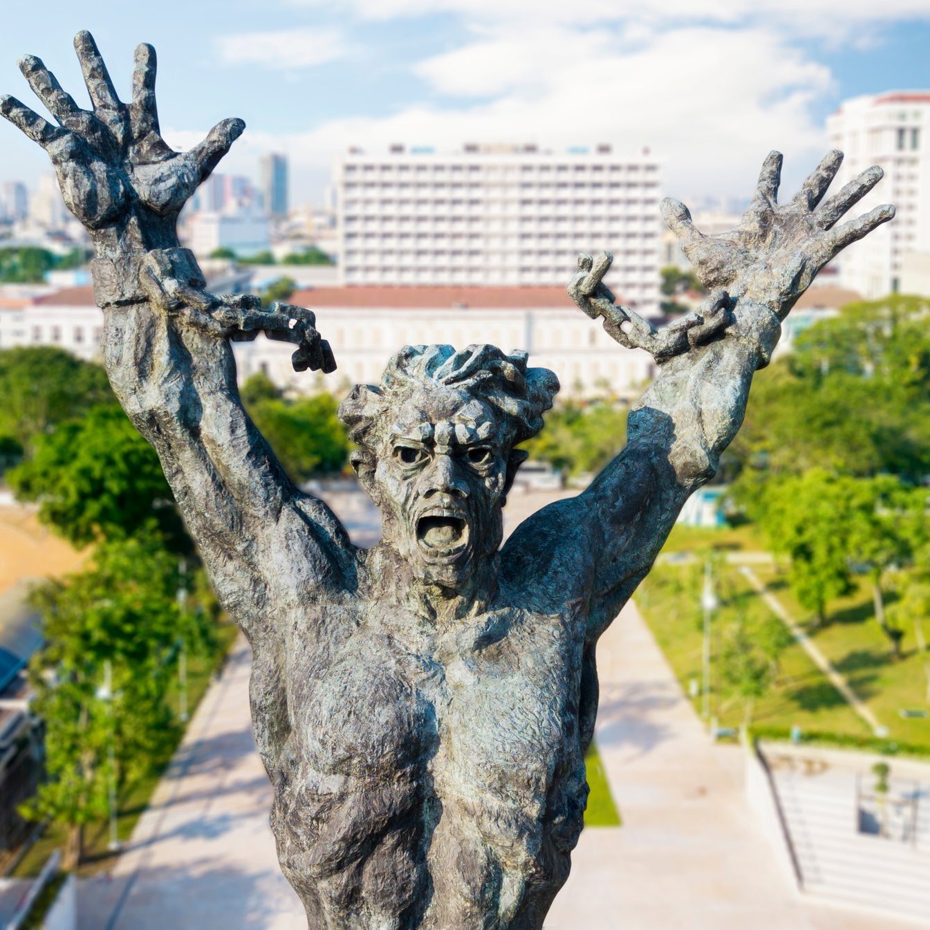 Cette image montre une statue impressionnante d'un homme, représenté dans une pose expressive. Il lève les bras vers le ciel, illustrant un sentiment intense, qui pourrait être celui de la lutte ou de l'angoisse. Son visage est déformé par une expression de détresse, accentuant une impression dramatique. La statue est en bronze, avec une texture riche et des détails finement sculptés qui soulignent les muscles et les traits faciaux. En arrière-plan, on aperçoit une ville moderne avec des bâtiments élevés et des espaces verts, créant un contraste entre la puissance de la sculpture et l'urbanisme environnant. Le ciel est dégagé, ajoutant une luminosité à la scène.