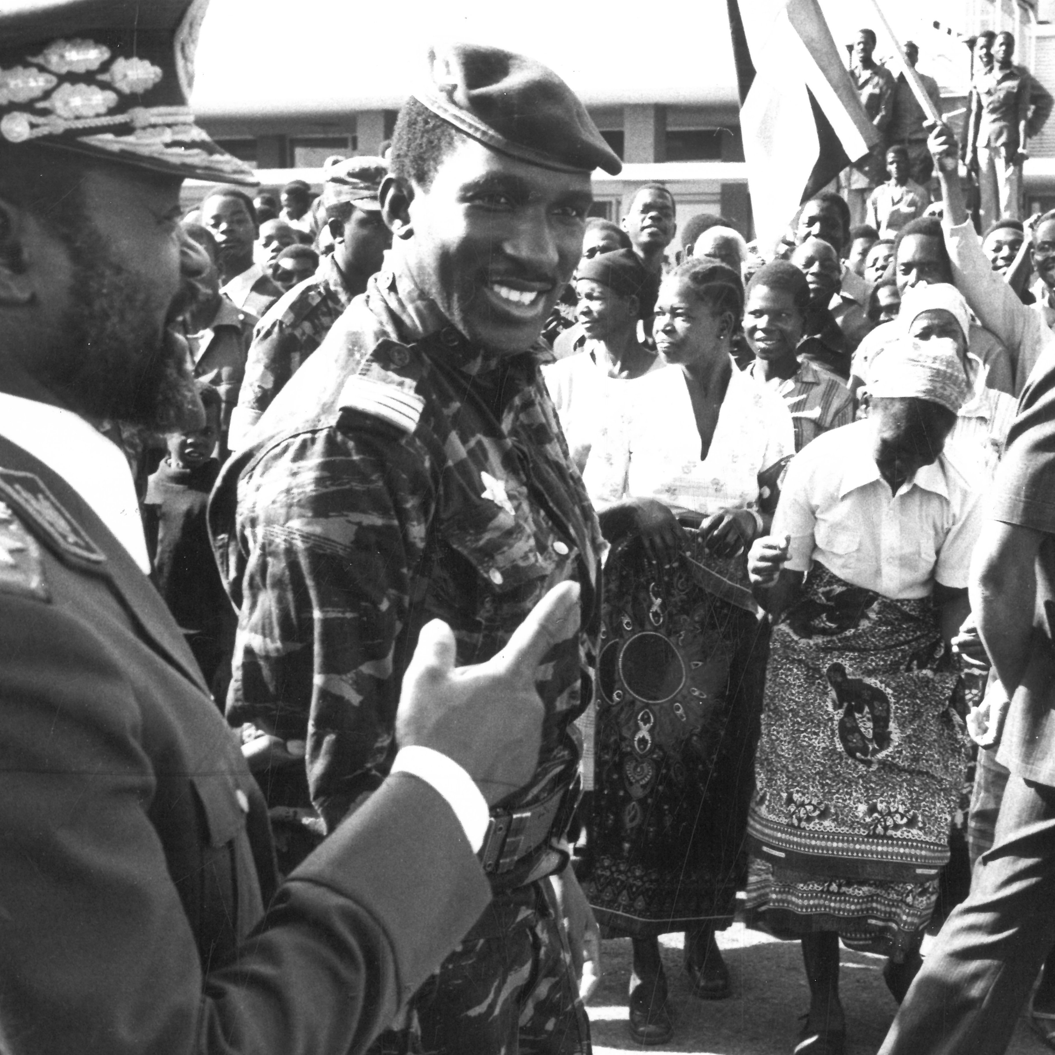 The image depicts a historical scene likely from a public event or rally. In the foreground, two men are engaging in conversation; one is dressed in a military uniform, while the other wears a military-style jacket with insignia. They both seem to be smiling and interacting positively. Behind them, a crowd of people of various ages is visible, with many holding flags and displaying enthusiastic gestures. The atmosphere appears lively, suggesting a moment of celebration or political significance. The image is in black and white, adding to its historical context.