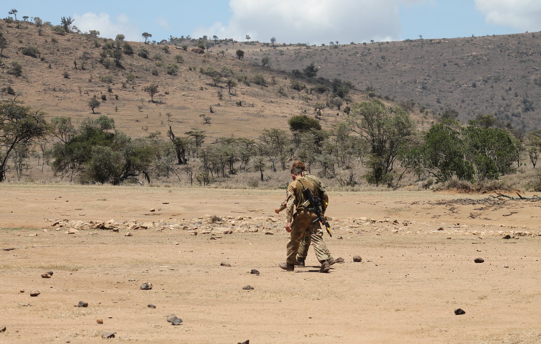 L'image montre un paysage aride et vaste, typique de la savane. Au centre, un homme marche seul sur un sol poussiéreux, parsemé de pierres. Il est vêtu d'un uniforme camouflage et porte un équipement à son dos, suggérant une activité en milieu naturel. En arrière-plan, on aperçoit des collines sèches et quelques arbres épars, témoignant d'une végétation clairsemée. Le ciel est partiellement nuageux, avec des nuages blancs qui contrastent avec le bleu du ciel. L'ensemble dégage une atmosphère de solitude et d'exploration dans un environnement sauvage.