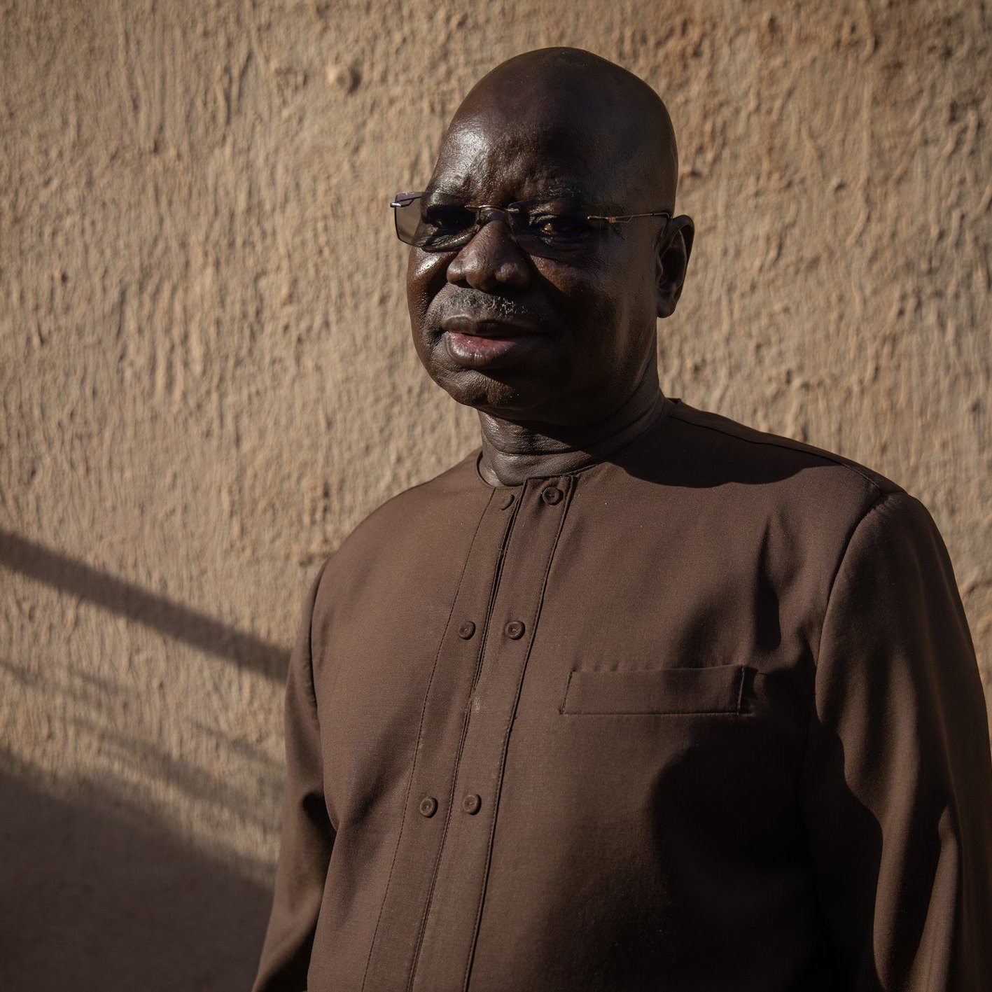 L'image présente un homme d'âge mûr, avec un teint foncé et des lunettes. Il porte une tenue sombre, légèrement scintillante, adaptée à un environnement formel ou culturel. Son expression est sereine et réfléchie. Le fond est constitué d'un mur en terre ou en adobe, éclairé par une lumière naturelle douce, créant une ambiance chaleureuse. Des ombres longues suggèrent qu'il est en fin de journée, ajoutant à la profondeur de l'image.
