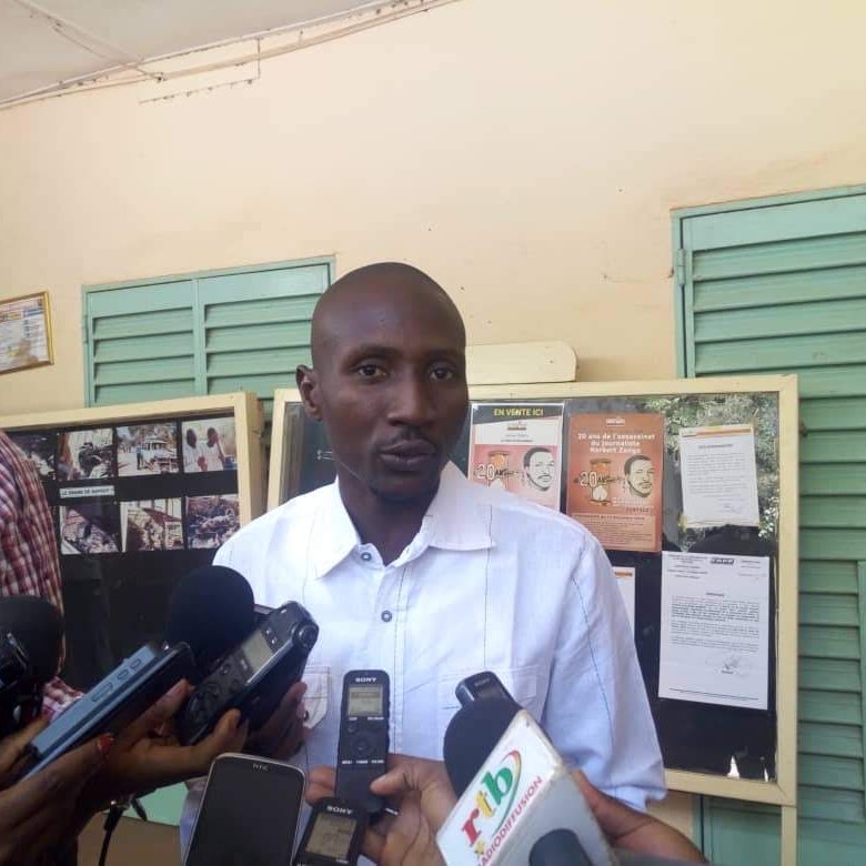 Dans cette image, un homme se tient devant un mur avec des panneaux d'affichage. Il porte une chemise blanche et semble parler à plusieurs journalistes qui l'entourent, tenant des microphones et des appareils d'enregistrement. Les panneaux d'affichage derrière lui contiennent des informations, des photos et des affiches. L'ambiance paraît être celle d'une conférence de presse ou d'un événement public où des informations sont partagées. 