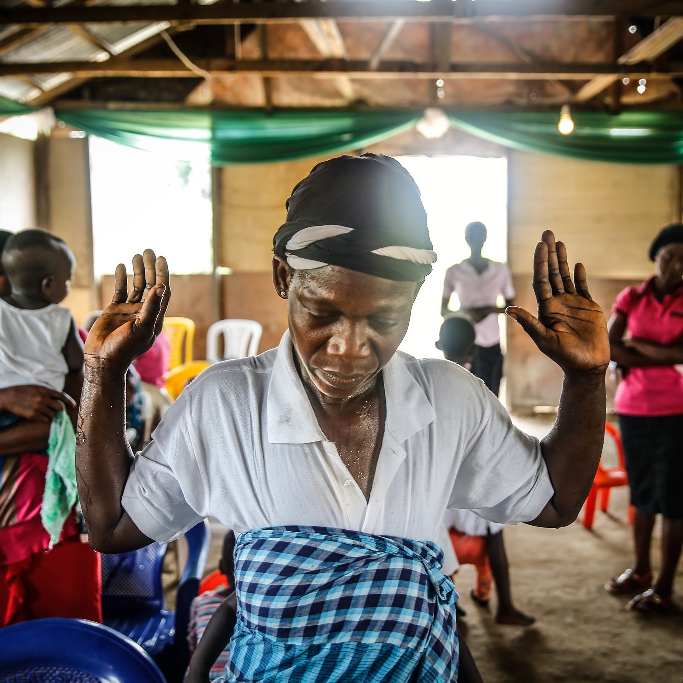 The image depicts a communal setting, likely a gathering or an event. In the foreground, a woman, dressed in a white top and a patterned shawl, raises her hands as if in prayer or meditation, demonstrating a moment of reverence or reflection. She appears focused and engaged in her spiritual practice. In the background, a variety of people are present, including individuals standing and others seated, indicating a diverse group participating in this activity. The interior of the space has wooden beams and a rustic charm, illuminated by soft lighting. The overall atmosphere seems to be one of community and spiritual connection.