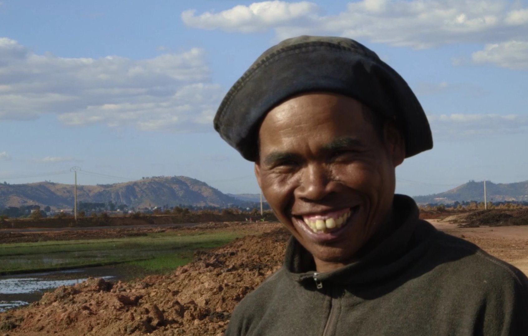 The image features a smiling man wearing a dark hat and a sweater. He is standing in an outdoor setting, likely near agricultural land, with a backdrop of hills and a partly cloudy sky. The ground appears to be bare earth, suggesting possible farming activity nearby. His cheerful expression conveys a sense of warmth and friendliness.