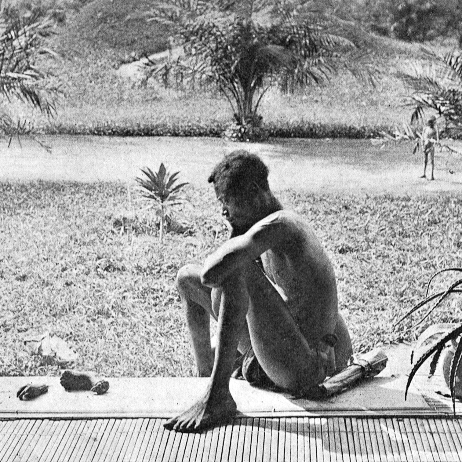L'image montre une scène en plein air, où une personne assise sur un rebord observe le paysage qui l'entoure. Ce personnage, de dos, semble pensif, en position accroupie, les jambes repliées. Il est nu et se trouve sur une surface en bois. À l'arrière-plan, il y a une étendue de verdure, avec des plantes tropicales comme des palmiers. Sur la gauche, deux enfants sont visibles, debout près d'un petit bosquet. Ils paraissent curieux, observant la scène. Au premier plan, on note des objets au sol, comme de petites pierres. L'atmosphère générale est tranquille et naturelle, évoquant un moment de réflexion dans un cadre sauvage.