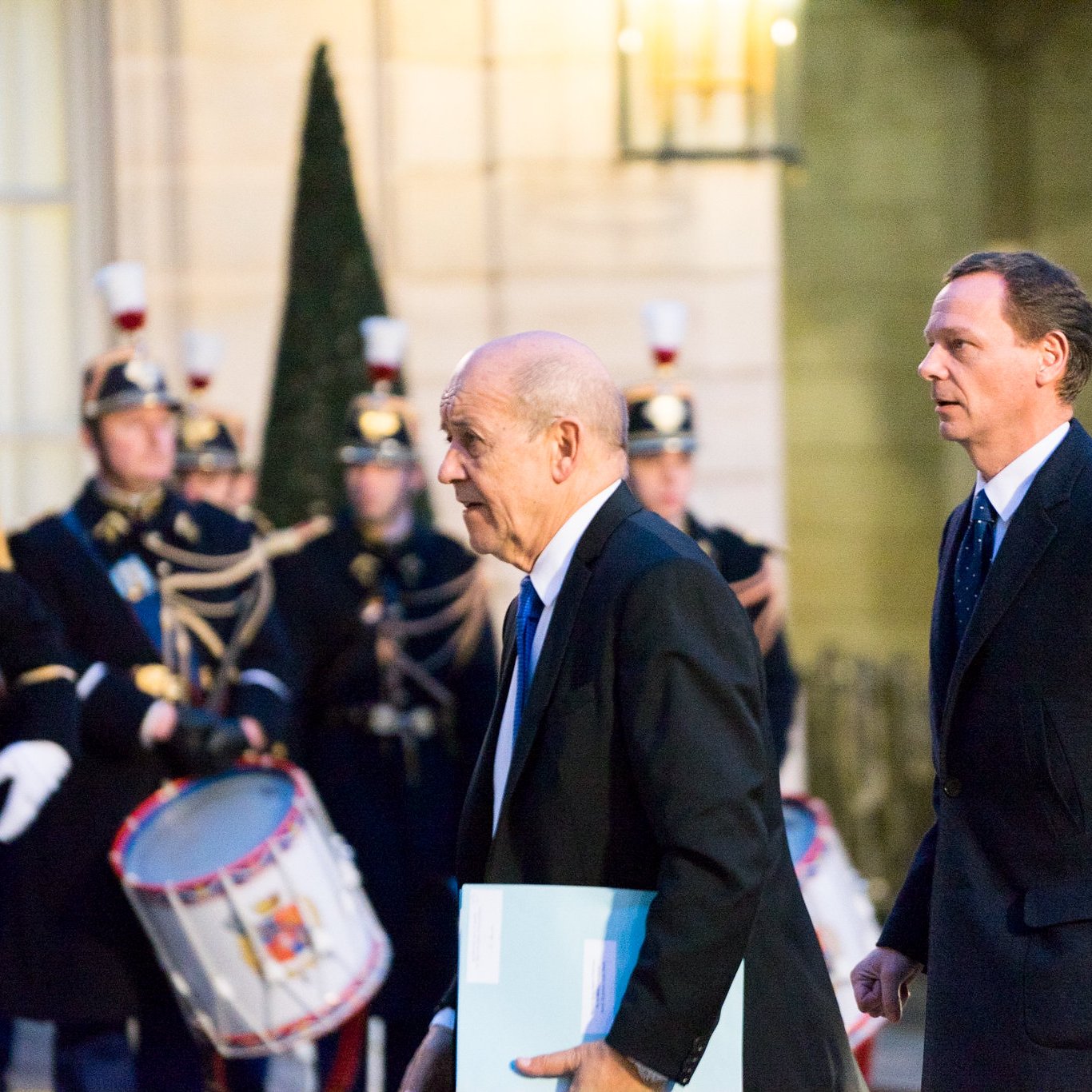 L'image montre deux hommes marchant dans une ambiance officielle. À l'arrière-plan, on peut apercevoir une garde cérémonielle, avec des soldats en uniforme traditionnel, portant des chapeaux distinctifs et tenant des instruments de musique, comme des tambours. Les hommes au premier plan semblent se rendre à une réunion importante, l'un d'eux portant un dossier ou un document. L'éclairage de la scène est doux, créant une atmosphère solennelle et respectueuse.