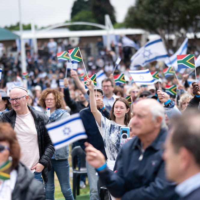 L'image montre une grande foule de personnes rassemblées dans un espace extérieur. Les gens tiennent des drapeaux, principalement des drapeaux israéliens, qui sont blancs et bleus avec l'Étoile de David, ainsi que des drapeaux sud-africains, qui présentent des couleurs vives. L'atmosphère est festive et engagée, avec des visages souriants et des expressions de joie. En arrière-plan, on peut apercevoir des assises et des personnes rassemblées autour d'un événement, suggérant une célébration ou une manifestation significative. Les vêtements des participants sont variés, allant de tenues décontractées à plus habillées, illustrant la diversité de la foule.