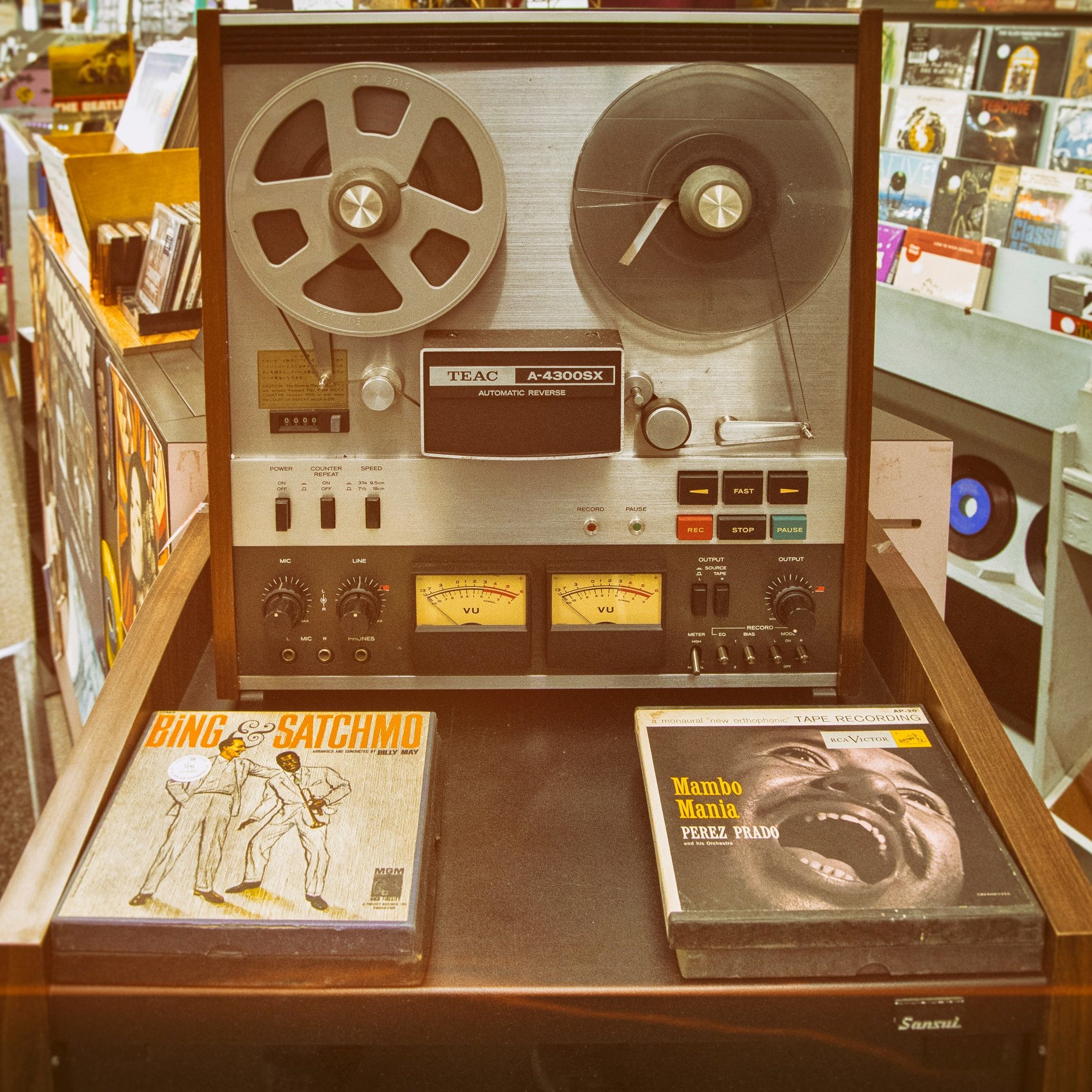 L'image montre un équipement audio vintage, un magnétophone à bande TEAC A-2300X, qui est posé sur un meuble en bois. Ce magnétophone a un design classique, avec un boîtier en métal argenté et des bobines de bande visibles sur le dessus. Deux compteurs analogiques sont situés en bas, indiquant les niveaux audio. À côté, on peut voir deux albums vinyles : l'un est intitulé "Bing & Satchmo", qui met en avant une collaboration entre le célèbre chanteur Bing Crosby et Louis Armstrong. L'autre album, "Mambo Mania" de Pérez Prado, suggère une atmosphère festive et rythmée. À l'arrière-plan, on aperçoit des étagères avec d'autres disques vinyles, créant une ambiance nostalgique et musicale.