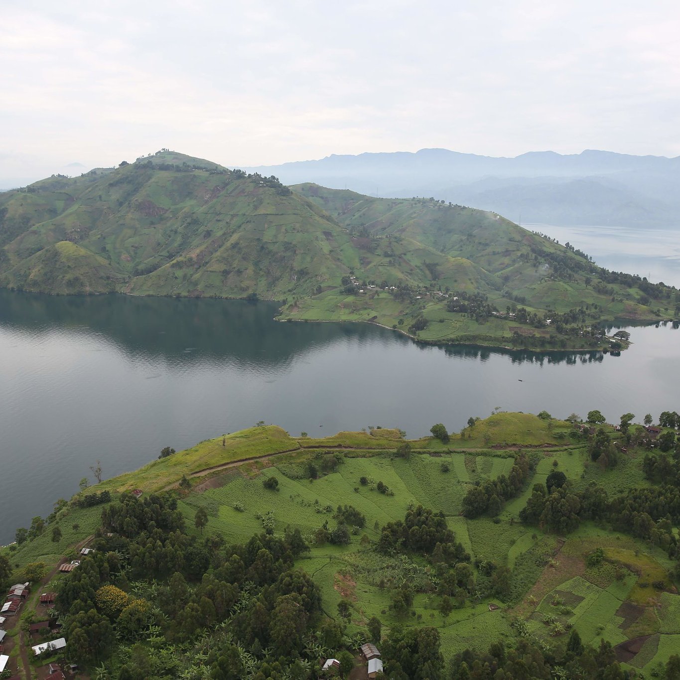 The image showcases a breathtaking landscape featuring lush green hills and a large lake. The hills are gently sloped and dotted with patches of vegetation, indicating a fertile area. In the foreground, there are scattered houses or structures, while the calm waters of the lake reflect the surrounding scenery. In the background, more mountainous terrain rises, adding depth to the view under a slightly overcast sky. The overall atmosphere is serene and picturesque, highlighting the natural beauty of the region.