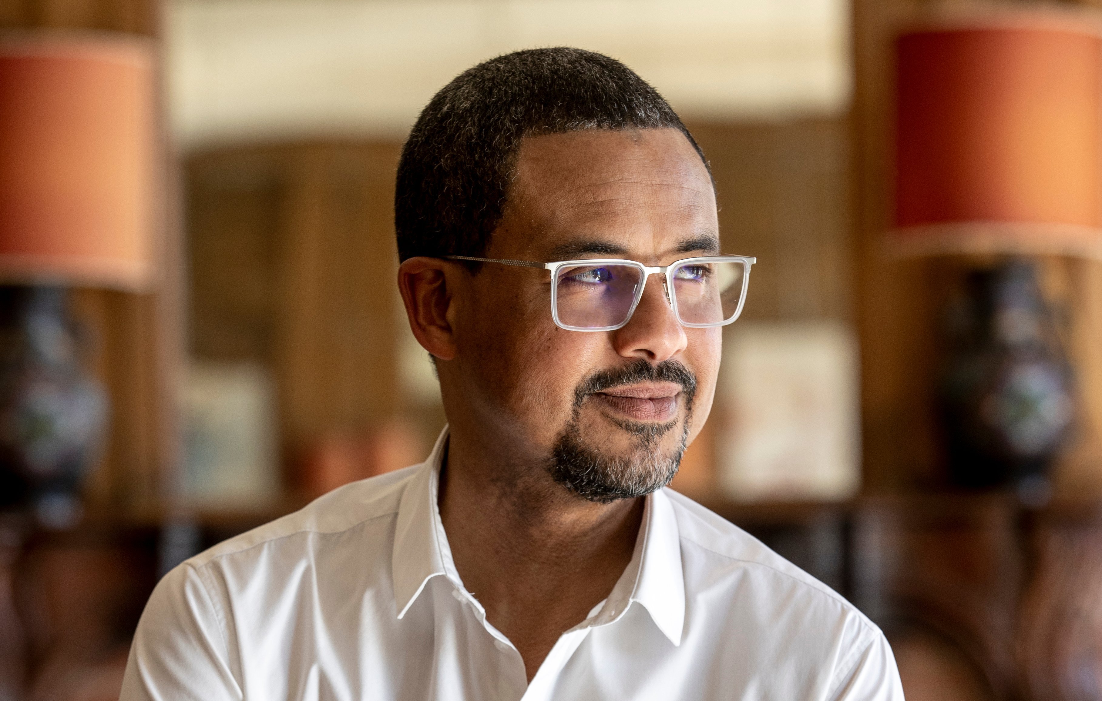 L'image présente un homme d'âge moyen, debout légèrement sur le côté. Il a la peau foncée et porte des lunettes avec une monture claire. Son expression est contemplative, avec un léger sourire aux lèvres. Il porte une chemise blanche, qui contraste avec son teint. En arrière-plan, on peut apercevoir des lampes avec des abat-jour orange et un décor en bois qui donne une ambiance chaleureuse et intime à l'ensemble. La lumière est douce, créant une atmosphère paisible.