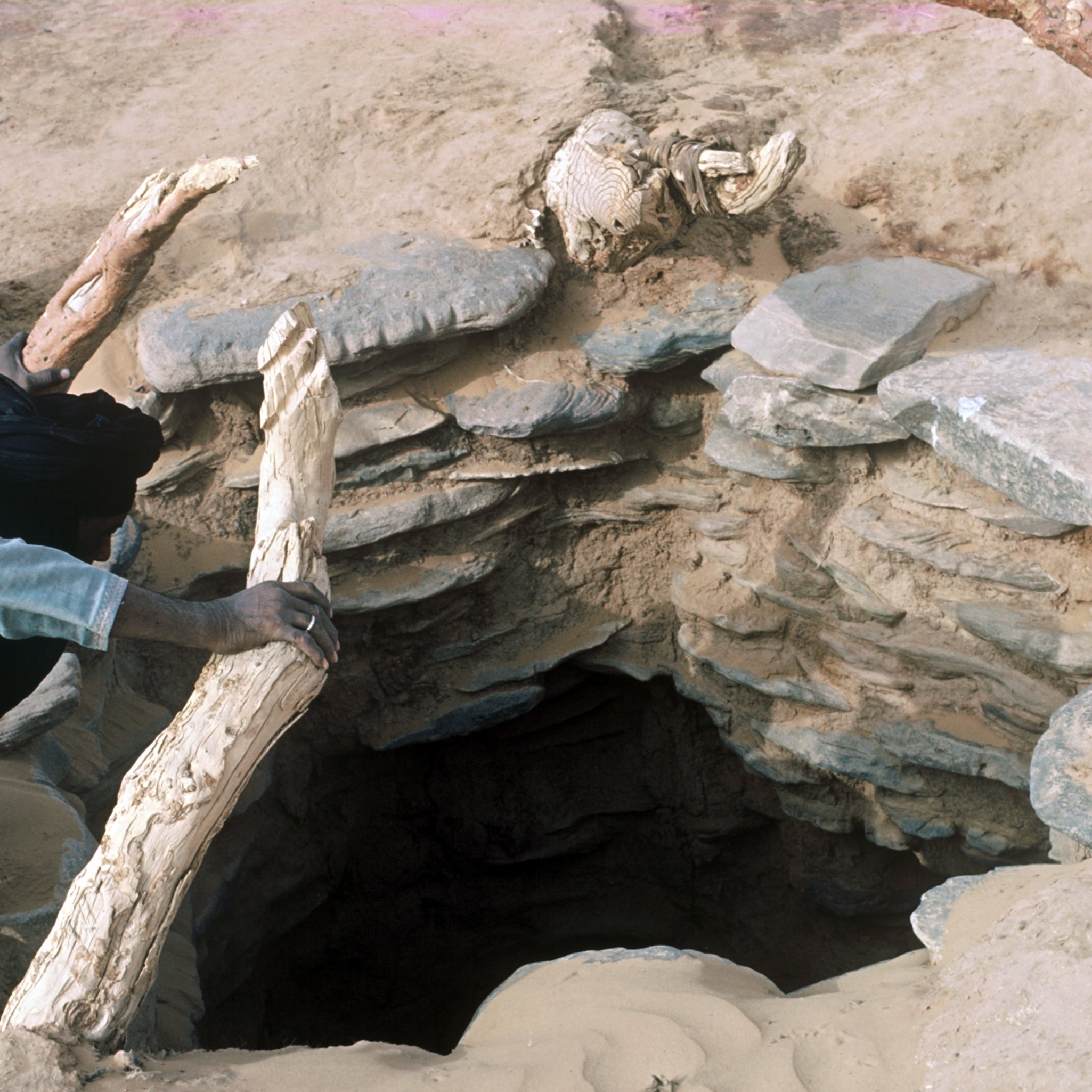 L'image montre un trou dans le sol, probablement un puits, entouré de pierres disposées de manière circulaire. À côté du trou, on aperçoit une personne, vêtue d'une chemise et d'un pantalon longs, qui semble s'approcher ou manipuler un morceau de bois. Le terrain est sablonneux, et des débris de bois sont visibles autour, suggérant une utilisation régulière de cet endroit. L'atmosphère semble aride, reflétant un environnement désertique.