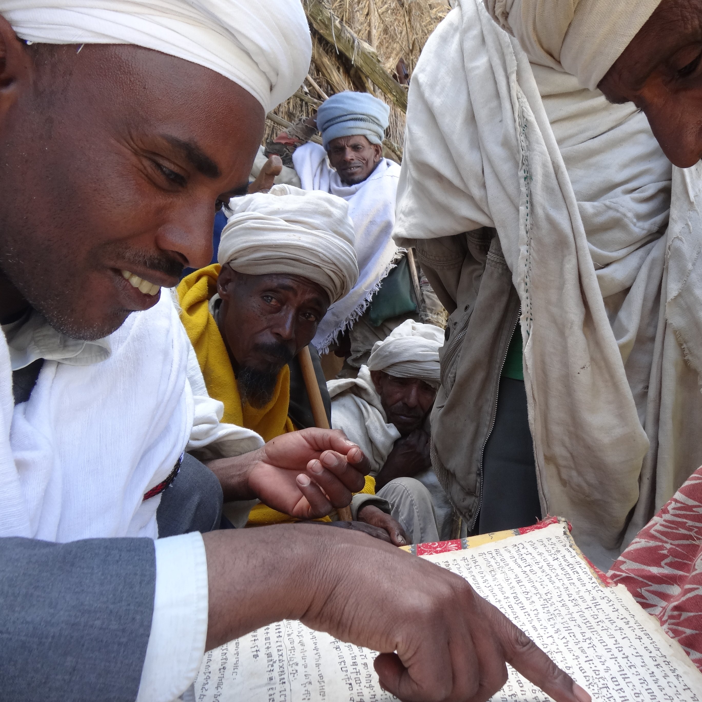 L'image montre un groupe de personnes, principalement des hommes, qui se rassemblent autour d'une table basse sur laquelle se trouve un ancien manuscrit. Un homme au premier plan, portant un habit traditionnel et un turban, montre du doigt une partie du texte. Les autres hommes, qui semblent intéressés, l'observent attentivement. Ils portent également des vêtements traditionnels, et leur expression trahit un mélange de curiosité et de respect. L'ambiance de la scène évoque un moment de partage et de transmission de connaissances, dans un cadre simple mais chargé de culture. En arrière-plan, des éléments naturels et des textures de tissus ajoutent une profondeur à l'atmosphère.