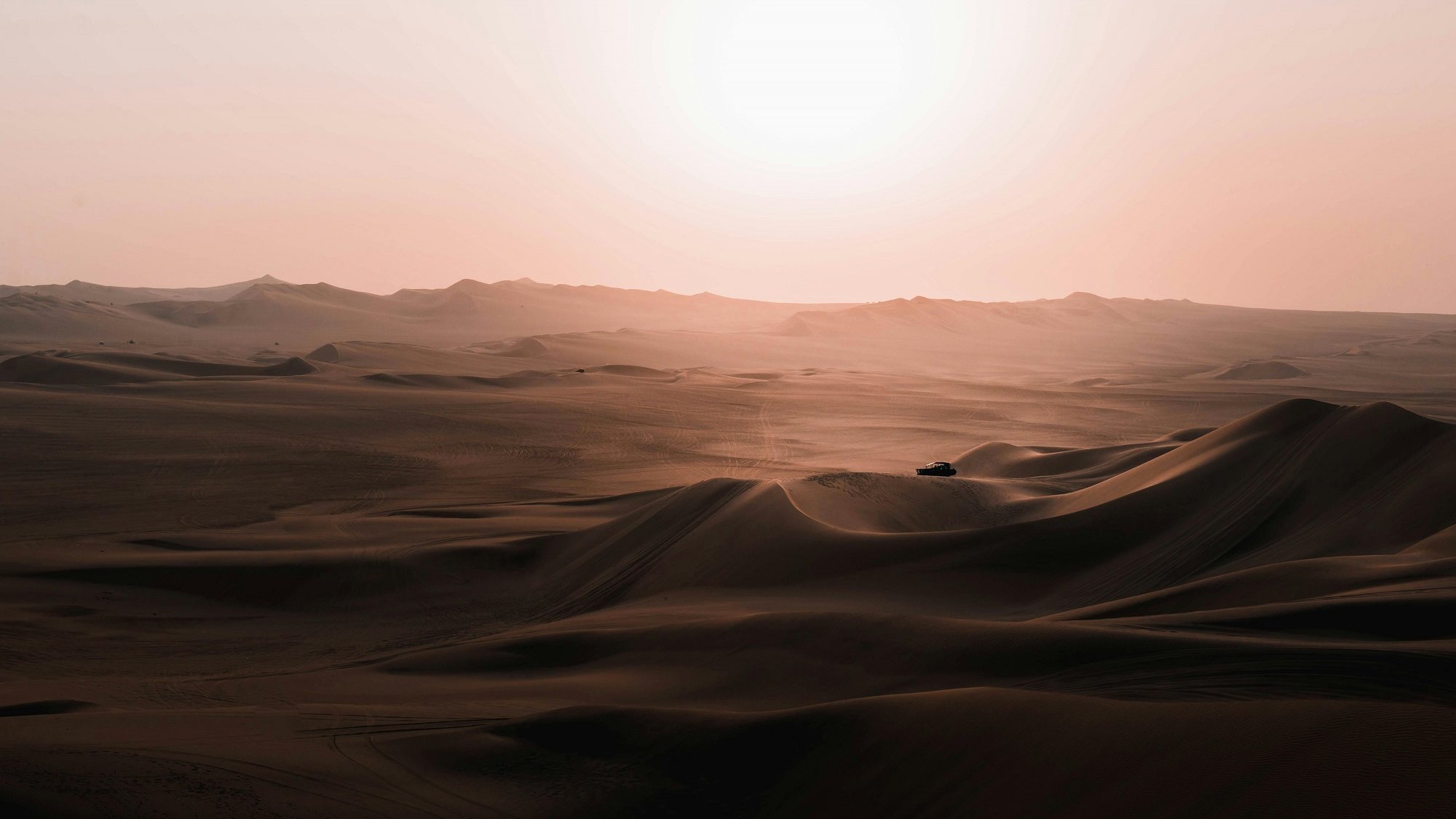 Imaginez une vaste étendue de sable, où les dunes s'étirent à perte de vue, leurs formes ondulantes créant des ombres douces sur le sol. Leurs contours se dessinent en vagues délicates, semblant danser sous les rayons d'un soleil flamboyant qui se lève à l'horizon, infusant le paysage d'une lumière chaude et dorée. Le silence règne, ponctué seulement par le léger murmure du vent qui balaie le désert, et une sensation de calme profonde imprègne l'air. Au loin, on devine une silhouette, peut-être un véhicule, qui semble minuscule face à l'immensité de ce panorama naturel. L'ensemble évoque une beauté brute, une tranquillité presque contemplative, comme si le temps s'était arrêté.