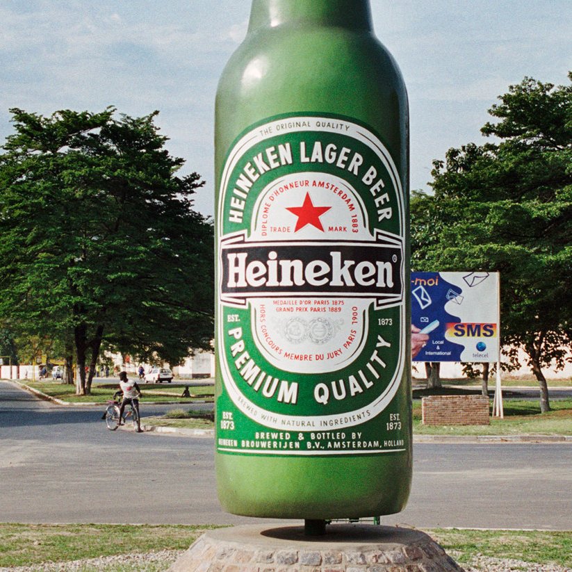 L'image montre une grande sculpture de bouteille de bière Heineken, qui se dresse au centre d'une intersection. La bouteille est peinte en vert vif et arborant l'étiquette rouge et blanche emblématique de la marque. Autour de la sculpture, on peut voir des gens se déplacer, certains à pied et d'autres à vélo. En arrière-plan, on distingue des arbres et quelques bâtiments qui ajoutent une ambiance locale. L'ensemble dégage une atmosphère animée et colorée, typique de l'urbanisme d'une ville vivante.