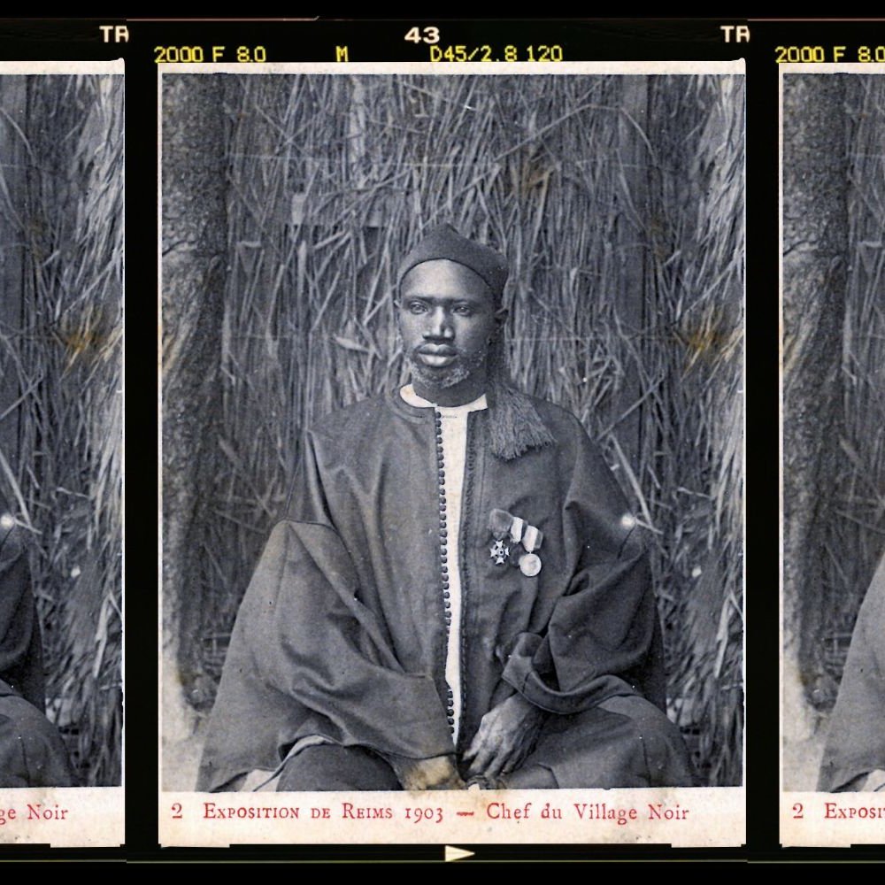 L'image présente trois portraits en noir et blanc d'un homme en costume traditionnel. Il est assis sur un fond de paille tressée qui évoque un environnement naturel. L'homme porte une tenue ample, probablement en textile sombre, et est décoré de plusieurs médailles accrochées à sa poitrine, ce qui suggère un statut élevé ou une reconnaissance. Son visage présente une expression solennelle et sérieuse. Les portraits sont disposés côte à côte, créant une composition symétrique. Cette image témoigne d'un moment historique, probablement lié à une exposition ou un événement marquant.