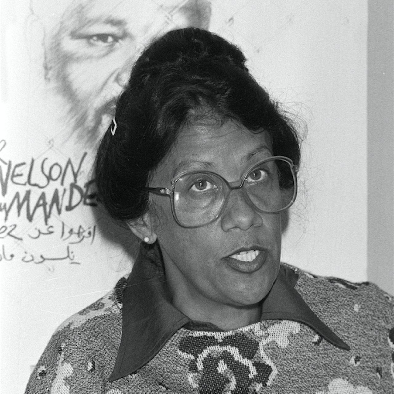 L'image montre une femme avec des lunettes épaisses, au visage expressif et aux cheveux noirs coiffés en un chignon. Elle porte un pull coloré avec un motif fleuri sur fond clair. En arrière-plan, on aperçoit une illustration d'un homme, probablement un portrait, qui ajoute un contexte à la scène. L'atmosphère semble sérieuse, et la femme semble engagée dans une discussion ou un discours. Ses traits et son regard transmettent une certaine détermination et une passion pour le sujet abordé.