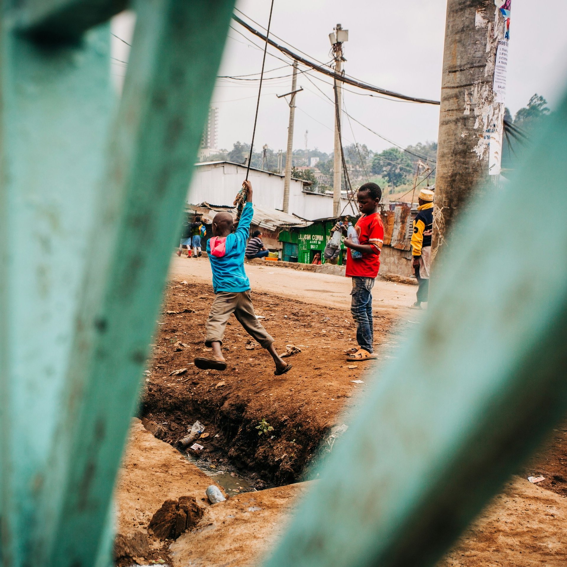 L'image présente une scène de la vie quotidienne dans un environnement urbain. Au premier plan, on aperçoit des barres d'une grille verte, qui encadrent la vue. À l'intérieur, deux enfants jouent près d'un chemin. L'un porte une chemise bleue et tient un objet au-dessus de sa tête, semblant s'amuser. L'autre enfant, aux vêtements rouges, observe ou se prépare à rejoindre le jeu. En arrière-plan, il y a des maisons simples, des poteaux électriques et une ambiance vibrante, typique d'un quartier animé. On sent une atmosphère de camaraderie et d'énergie enfantine. Le terrain est boueux et légèrement en pente.