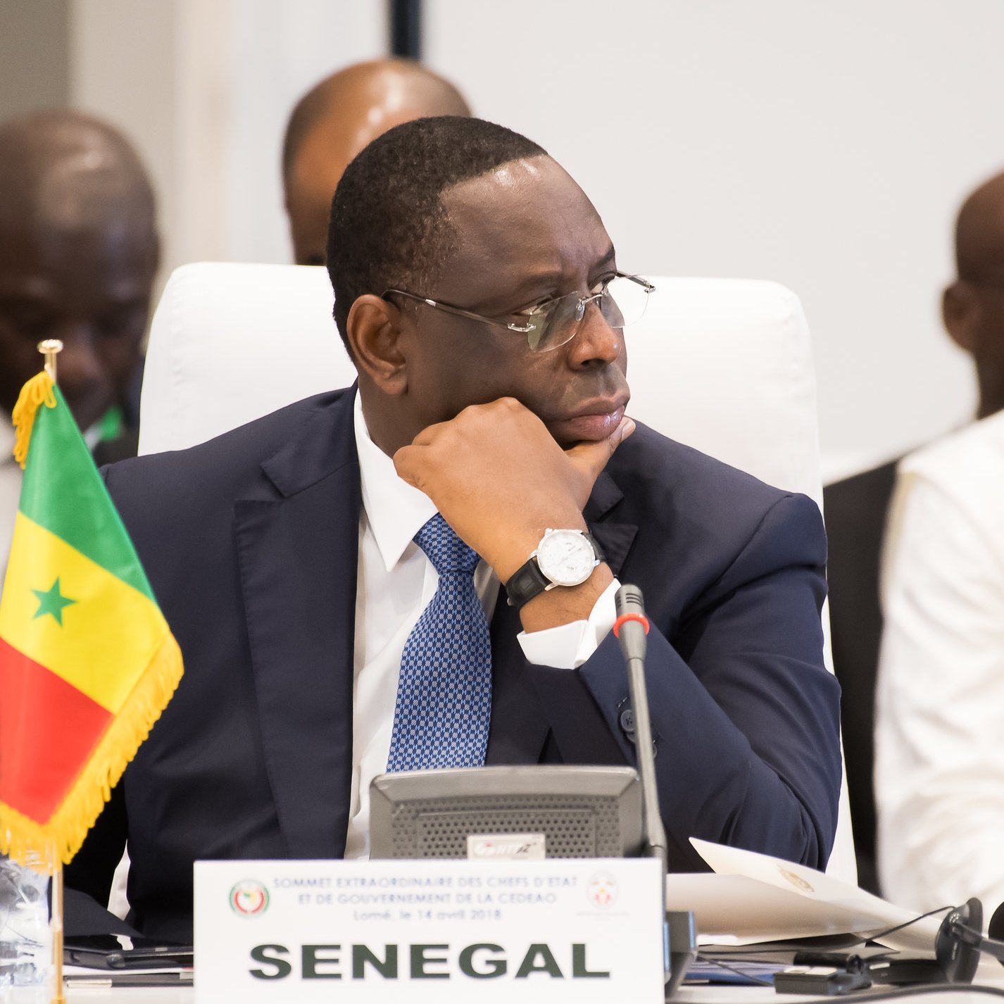 L'image montre un homme assis à une table dans un cadre formel. Il porte un costume sombre et un bracelet montre argenté. Son visage exprime une concentration sérieuse, alors qu'il regarde pensivement devant lui. À sa gauche, il y a un drapeau du Sénégal planté sur la table, indiquant son origine ou son rôle. En arrière-plan, d'autres personnes sont présentes, mais ne sont pas clairement visibles. L'ambiance de la scène suggère un événement officiel, peut-être une réunion ou une conférence de dirigeants.