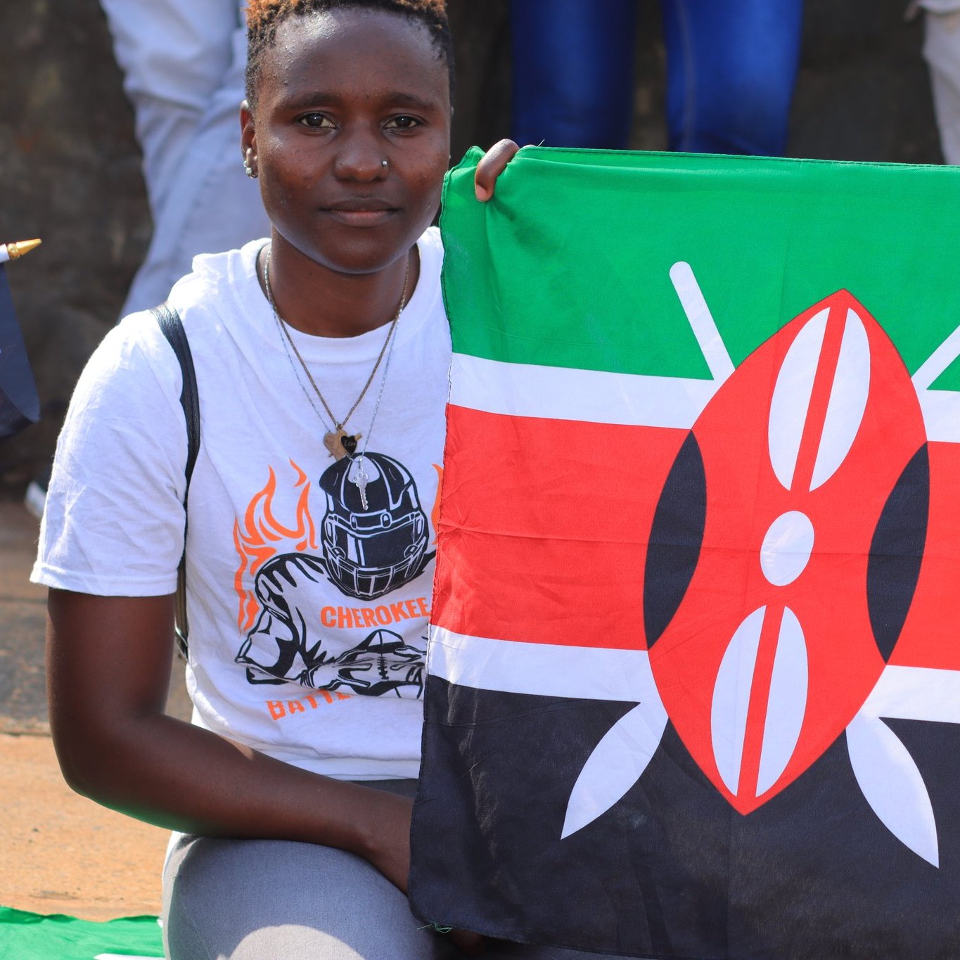 Dans cette image, une jeune femme sourit tout en tenant une grande drapeau du Kenya. Le drapeau est composé de bandes horizontales de couleur noire, rouge et verte, avec un motif central représentant un écu et des flèches. La femme porte un t-shirt blanc orné d'un motif sportif, avec un choker en pendentif autour du cou. En arrière-plan, on peut apercevoir d'autres personnes, certaines tenant également des drapeaux, créant une ambiance festive et patriotique.