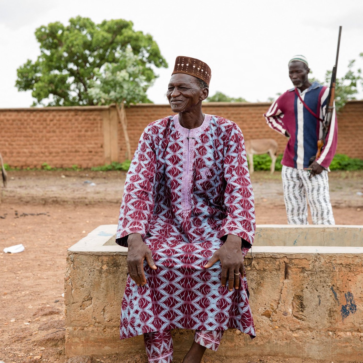 L'image montre un homme assis sur un mur en béton dans un environnement rural. Il porte une tenue traditionnelle, ornée de motifs géométriques dans des tons de rouge et blanc, et un chapeau conique typique. Son expression est détendue et souriante, suggérant une ambiance joyeuse. À l'arrière-plan, un deuxième homme se tient debout, avec un fusil à la main, veillant sur des ânes qui paissent paisiblement. La scène évoque une vie simple et traditionnelle, ancrée dans la nature. Le ciel est nuageux, ajoutant une légère atmosphère de sérénité au paysage.