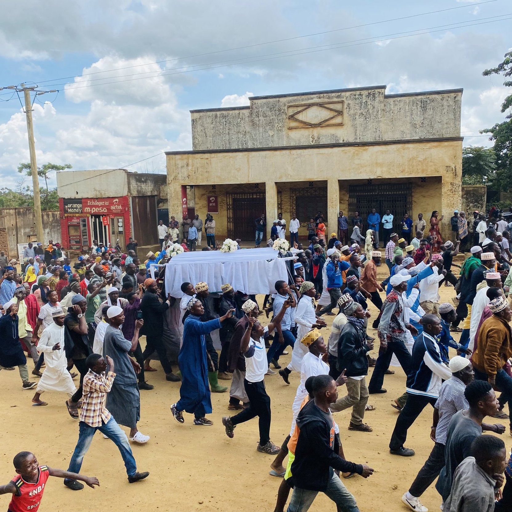 L'image montre une scène animée dans une rue où une grande foule se déplace. Au centre de l'attention, des personnes portent un drap blanc qui semble recouvrir un cercueil, indiquant qu'il s'agit d'un cortège funéraire. Les membres de la foule sont vêtus de divers habits, certains en tenues traditionnelles, d'autres plus contemporaines. Les visages reflètent des émotions variées, allant du recueillement à la tristesse. À l'arrière-plan, on peut remarquer des bâtiments aux murs fissurés, avec des panneaux publicitaires visibles. Le ciel est légèrement nuageux, créant une atmosphère à la fois sombre et lugubre, mais la scène reste vibrante de vie grâce à la présence de nombreux participants.
