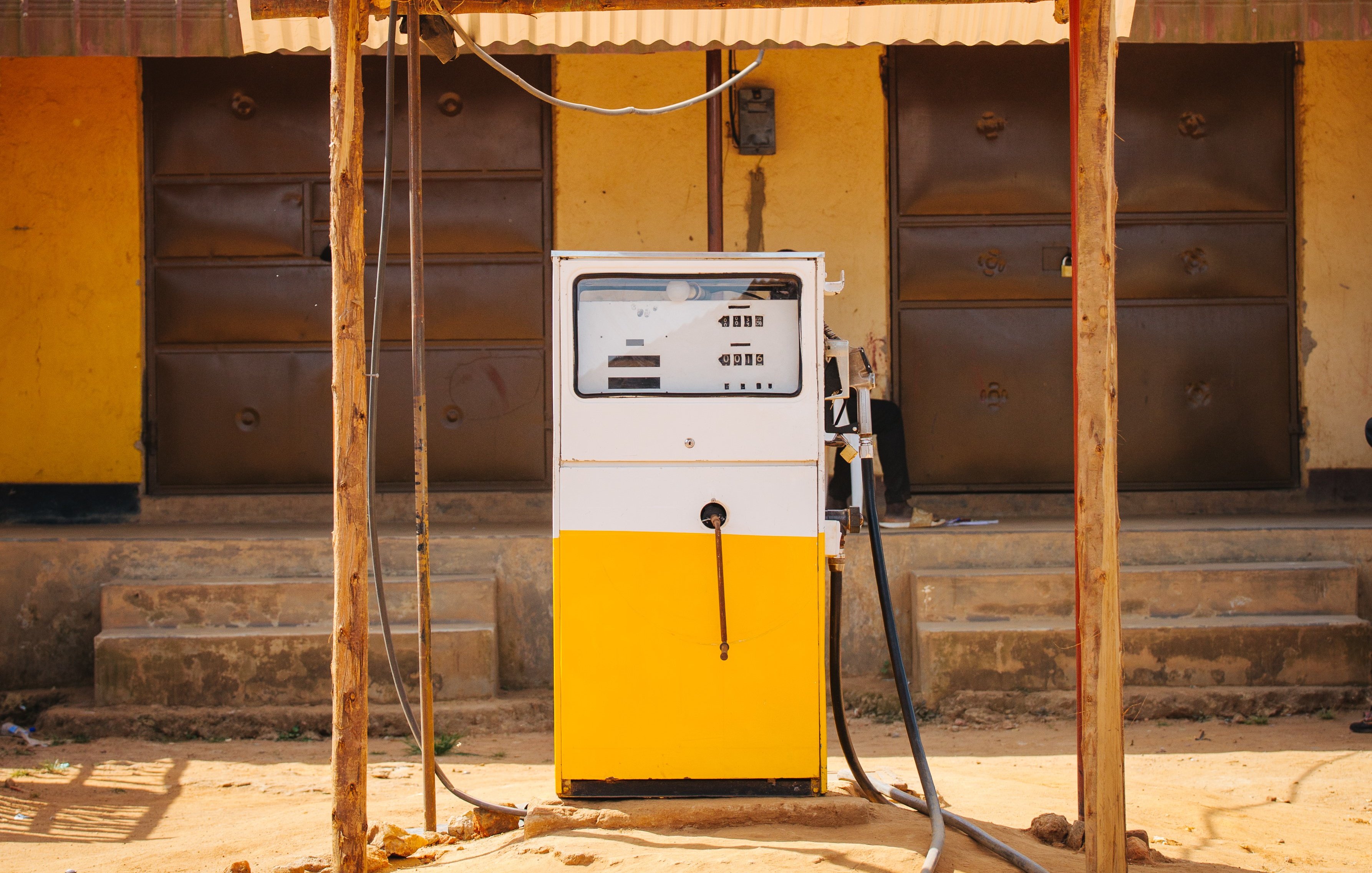 Imaginez une petite station-service située à l'extérieur d'un bâtiment. Au centre de l'image se trouve une pompe à essence de couleur blanche et jaune, installée sous un auvent en tôle ondulée qui offre un peu d'ombre. La pompe est équipée d'un écran et d'un bouton pour le paiement, ainsi qu'un flexible noir reliant la pompe au réservoir d'un véhicule. À l'arrière-plan, on aperçoit un mur de couleur jaune avec deux portes en métal marron, fermées, et l'ensemble est posé sur un sol terreux, suggérant une ambiance rustique et simple. Le lieu semble tranquille, sans voitures visibles autour de la pompe.