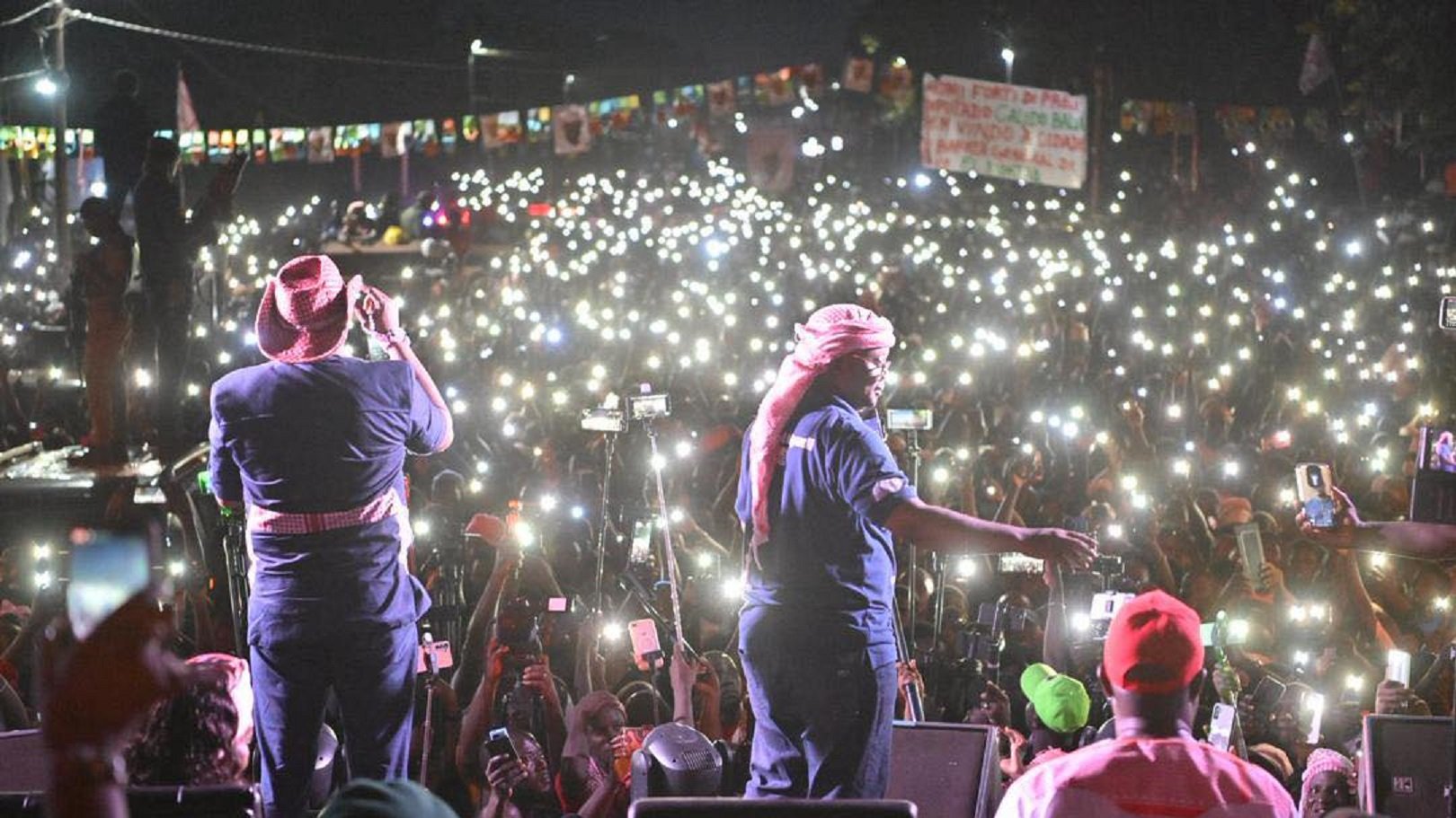 L'image montre une scène d'un concert ou d'un rassemblement en plein air, où un grand nombre de personnes est rassemblé devant un podium. Au premier plan, deux artistes sont sur scène, l'un chantant et l'autre en mouvement, tous deux vêtus de vêtements décontractés, avec des chapeaux et des bandanas. Derrière eux, une mer de spectateurs se tient en arrière-plan, illuminée par d'innombrables lumières de téléphones portables levés, créant un effet scintillant dans la nuit. L'atmosphère est électrisante, avec des couleurs vives provenant des vêtements des membres du public et des affiches. Des banderoles et des drapeaux sont également visibles, ajoutant à l'ambiance festive et engagée de l'événement.