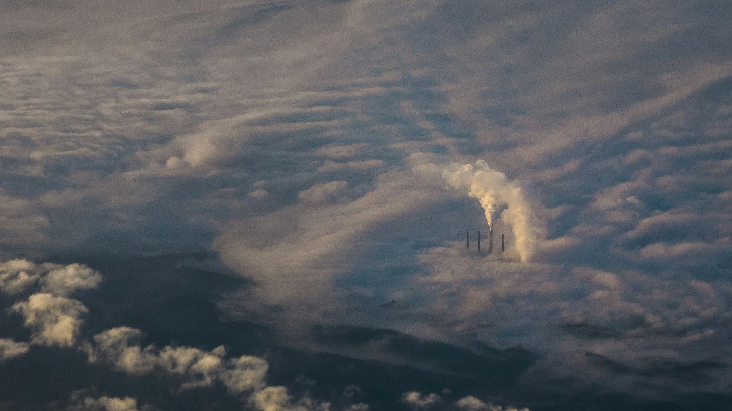 L'image présente une vue aérienne d'un paysage nuageux. On peut distinguer plusieurs cheminées d'une centrale, émettant des colonnes de fumée blanche qui se fondent dans les nuages environnants. Les nuages eux-mêmes paraissent denses et vaporeux, créant une atmosphère mystique. La lumière semble filtrer à travers le brouillard, ajoutant une touche douce au paysage. Dans l'ensemble, l'image évoque une impression de grandeur et de puissance, avec la nature et l'industrialisation coexistant dans un environnement nuageux.
