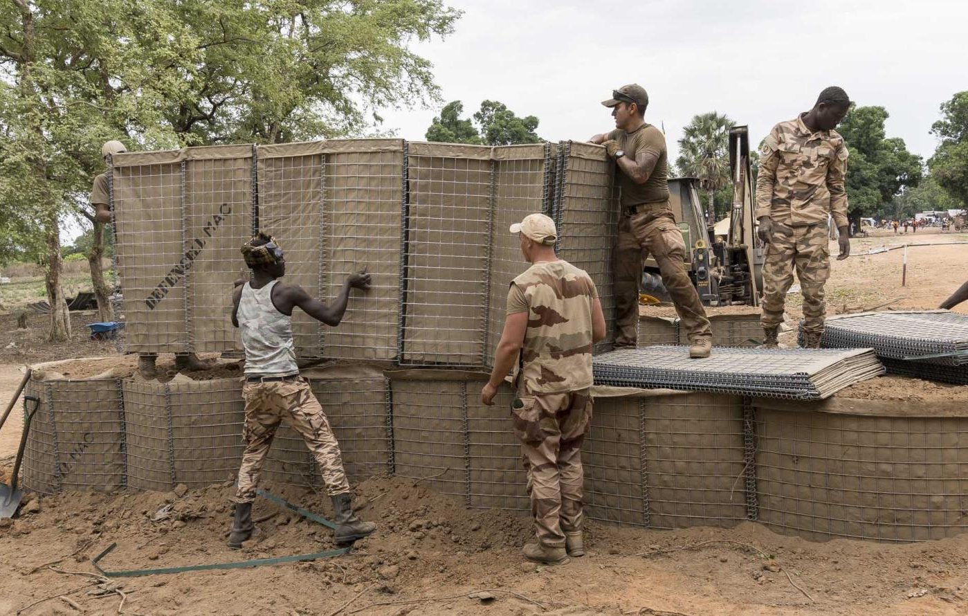L'image montre un groupe de soldats au travail sur un site de construction. Ils sont en train de manipuler de grandes structures en treillis, qui semblent être remplies de terre. L'environnement est naturel, avec des arbres verts en arrière-plan et un sol légèrement argileux. Les soldats portent des uniformes militaires et semblent concentrés sur leur tâche. Certains d'entre eux soulèvent des éléments, tandis que d'autres les positionnent soigneusement. Cette scène évoque une atmosphère de collaboration et d'effort collectif.