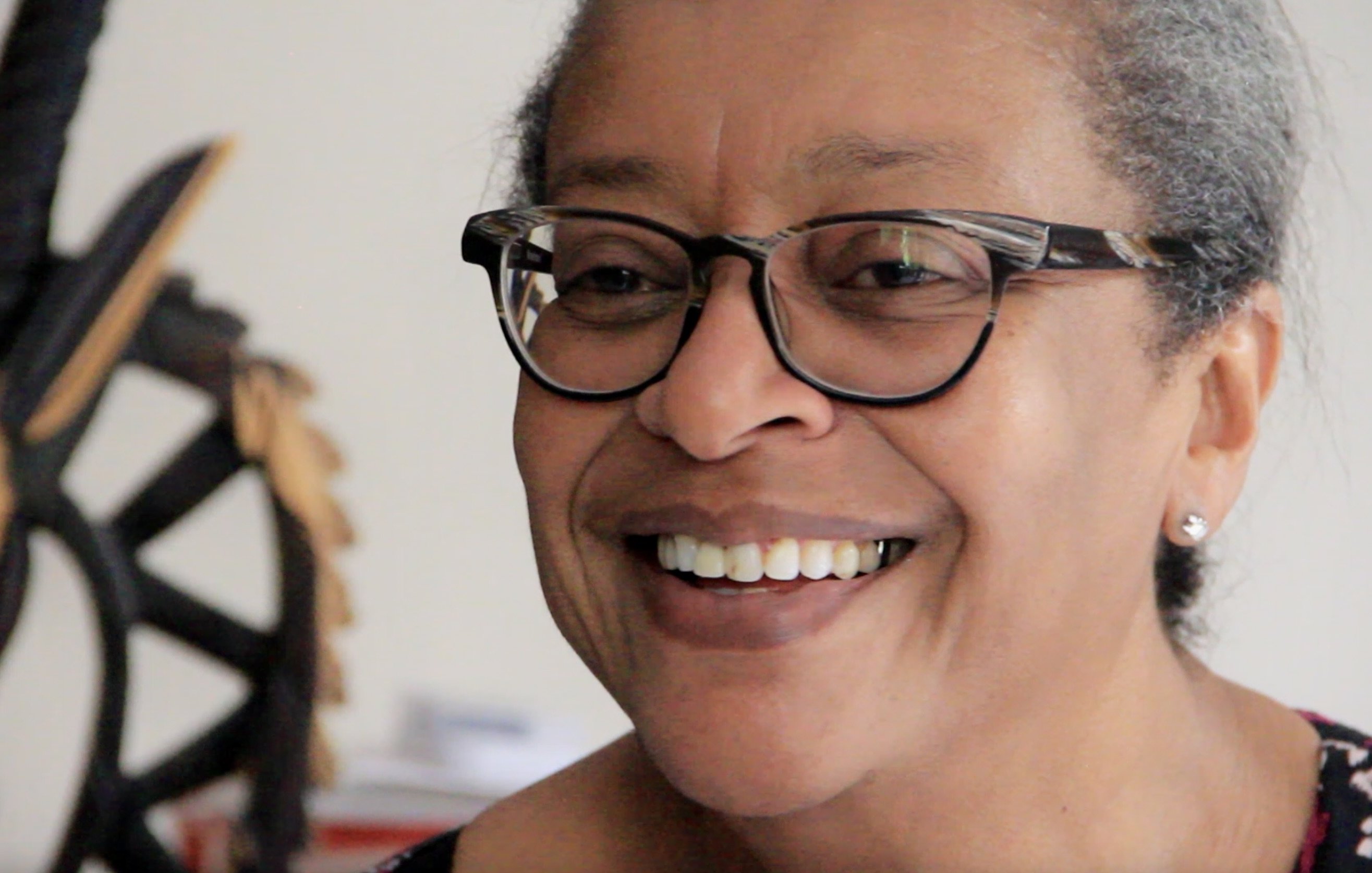 L'image montre une femme souriante, avec des lunettes à monture noire. Son expression est chaleureuse et accueillante. Elle a les cheveux grisonnants, peignés en arrière, et porte des boucles d'oreilles discrètes. En arrière-plan, on aperçoit une sculpture en bois, qui semble représenter un motif stylisé. L'atmosphère est conviviale et intime, suggérant un moment de bonheur ou de partage.