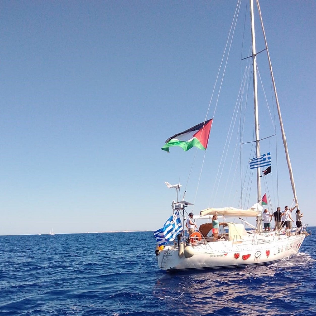 L'image présente un voilier sur une mer turquoise calme, sous un ciel d'un bleu éclatant sans nuages. Le bateau, de couleur blanche, arbore plusieurs drapeaux, dont celui de la Palestine, de la Grèce, et d'autres drapeaux en bandes horizontales typiques. À bord, on aperçoit des personnes qui semblent profiter d'une journée en mer. Les vagues légères créent une ambiance paisible, et l'horizon lointain s'étend, avec d'autres bateaux visibles au loin. L'ensemble évoque une atmosphère de liberté et d'aventure maritime.