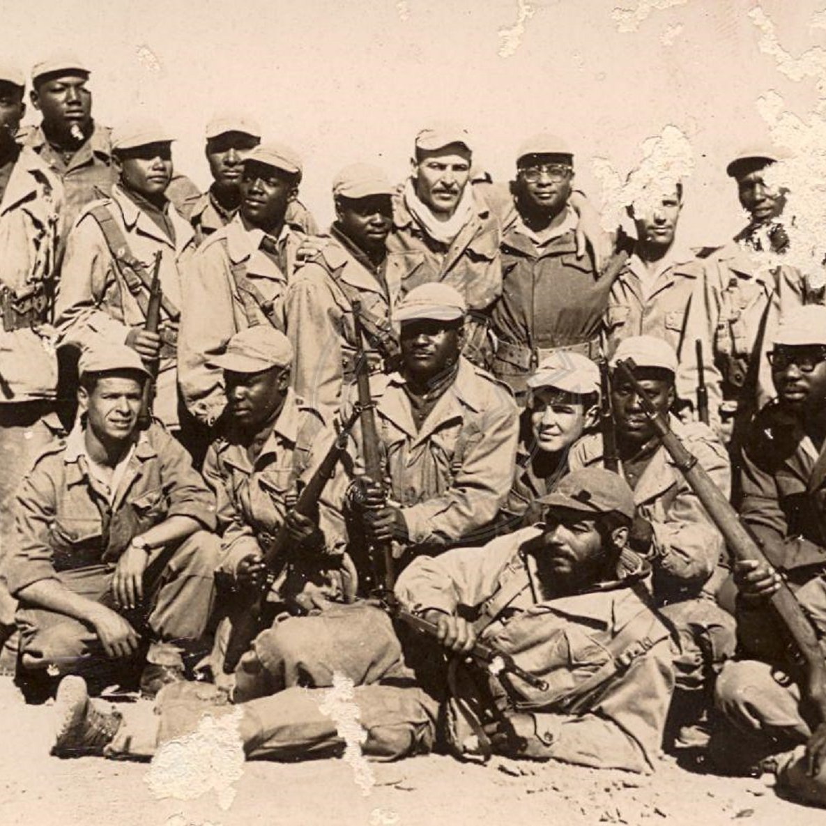 Cette image montre un groupe de soldats, probablement dans un contexte historique. Ils sont vêtus d'uniformes militaires, la plupart portant des chapeaux ou casquettes. Certains d'entre eux tiennent des armes, et l'ambiance générale dégage un sentiment de camaraderie et de force. Les soldats semblent être regroupés, avec une variation dans leurs postures : certains se tiennent debout tandis que d'autres sont accroupis ou assis. Le fond de la photo est déteint, ce qui suggère qu'il pourrait s'agir d'une image ancienne. L'ensemble de cette scène évoque un moment de solidarité et de détermination au sein du groupe.