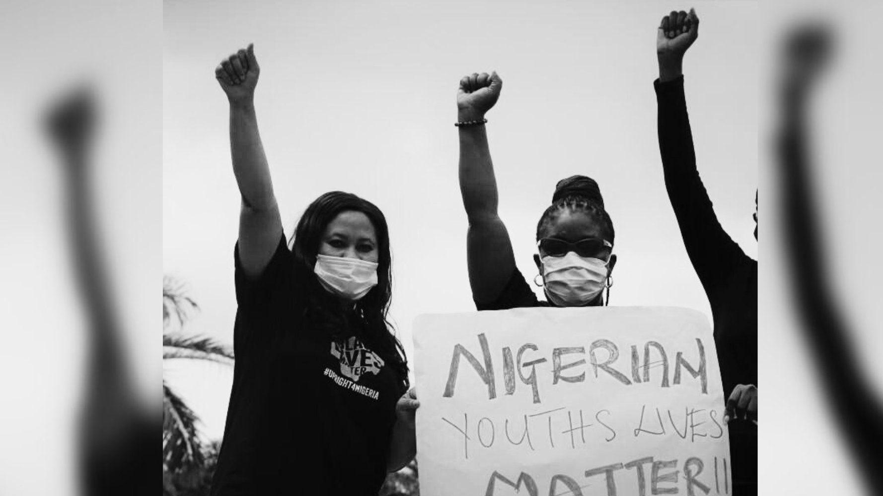L'image montre un groupe de personnes participant à une manifestation. Deux femmes, portant des masques, lèvent le poing en signe de solidarité. Elles affichent des expressions déterminées. L'une d'elles tient une pancarte sur laquelle il est écrit "NIGERIAN YOUTHS LIVES MATTER !!". L'arrière-plan est flou, mais on peut imaginer un espace extérieur, suggérant une atmosphère de mobilisation et de revendication. L'absence de couleurs, avec un effet en noir et blanc, accentue le sérieux et l'urgence du message porté par les manifestants.