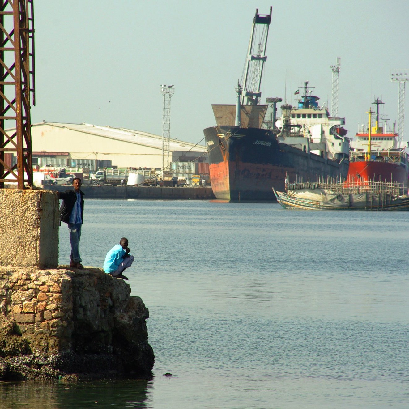 Imaginez un port animé. Au premier plan, il y a un quai en pierre avec plusieurs personnes : certaines sont debout, discutant, tandis que d'autres se penchent sur le bord pour observer l'eau. L'atmosphère est vivante, remplie d'échos de rires et de conversations. À l'arrière-plan, de grands navires sont amarrés, leurs coques visibles au-dessus de l'eau paisible. Des grues et des poteaux élancés se dressent, témoignant des activités portuaires. L'air sent légèrement le sel et le poisson, créant une ambiance maritime authentique.